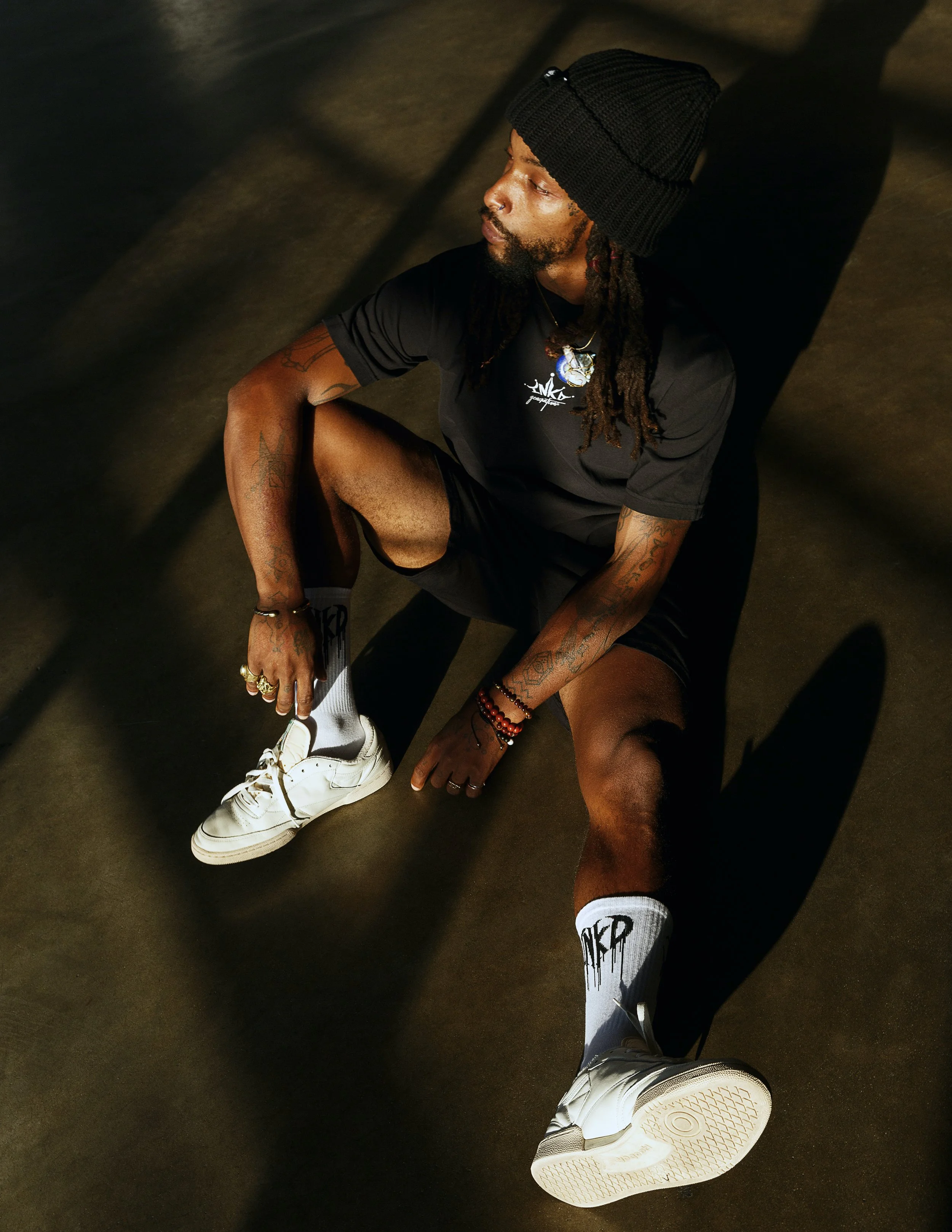 A person with dreadlocks, tattoos, and jewelry sitting on the ground with one knee up, wearing a black beanie, black T-shirt, white sneakers, and white socks, with shadows cast on the floor.