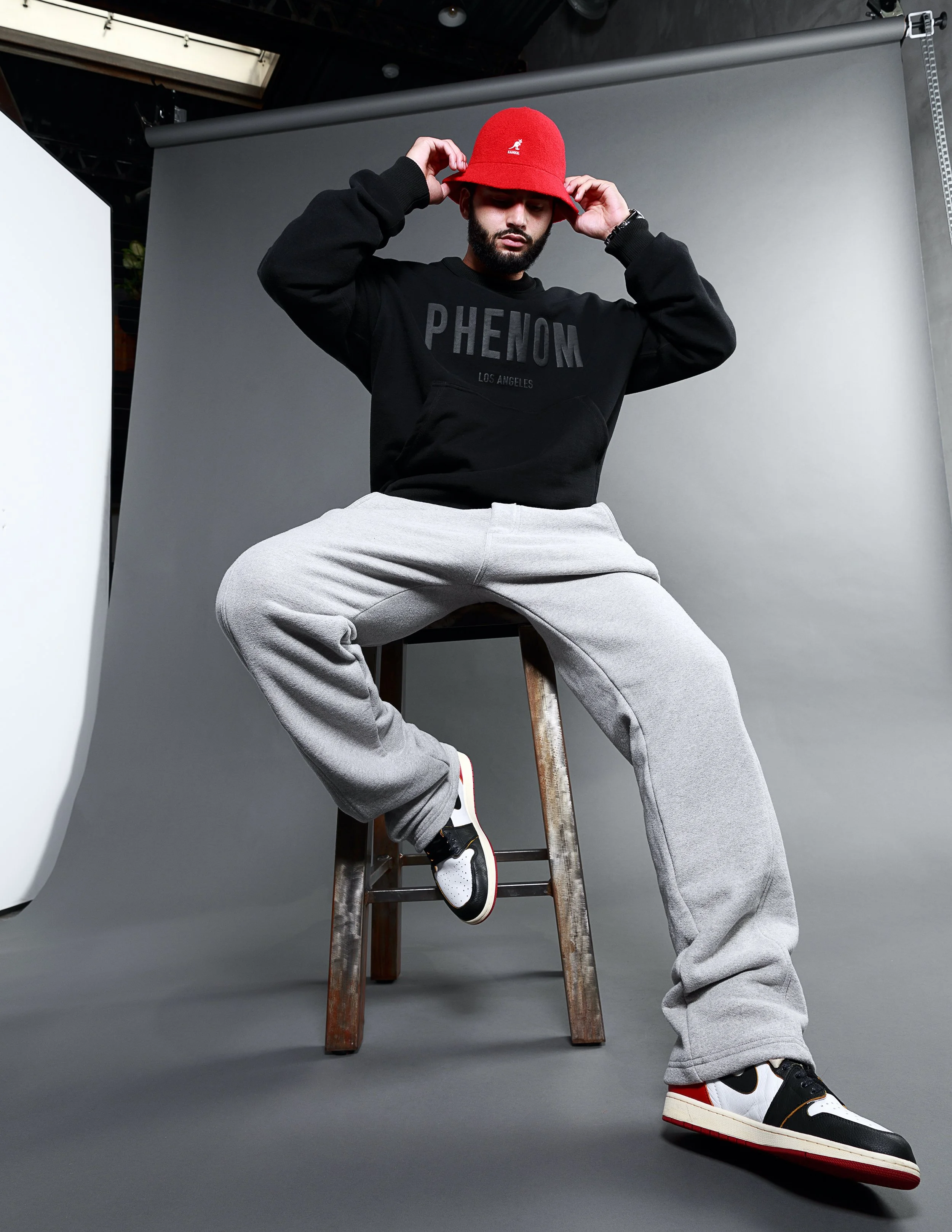 A man wearing a red bucket hat, black sweatshirt with 'PHENOM LOS ANGELES' written on it, gray sweatpants, and black, white, and red sneakers, sitting on a wooden stool against a gray background in a studio.