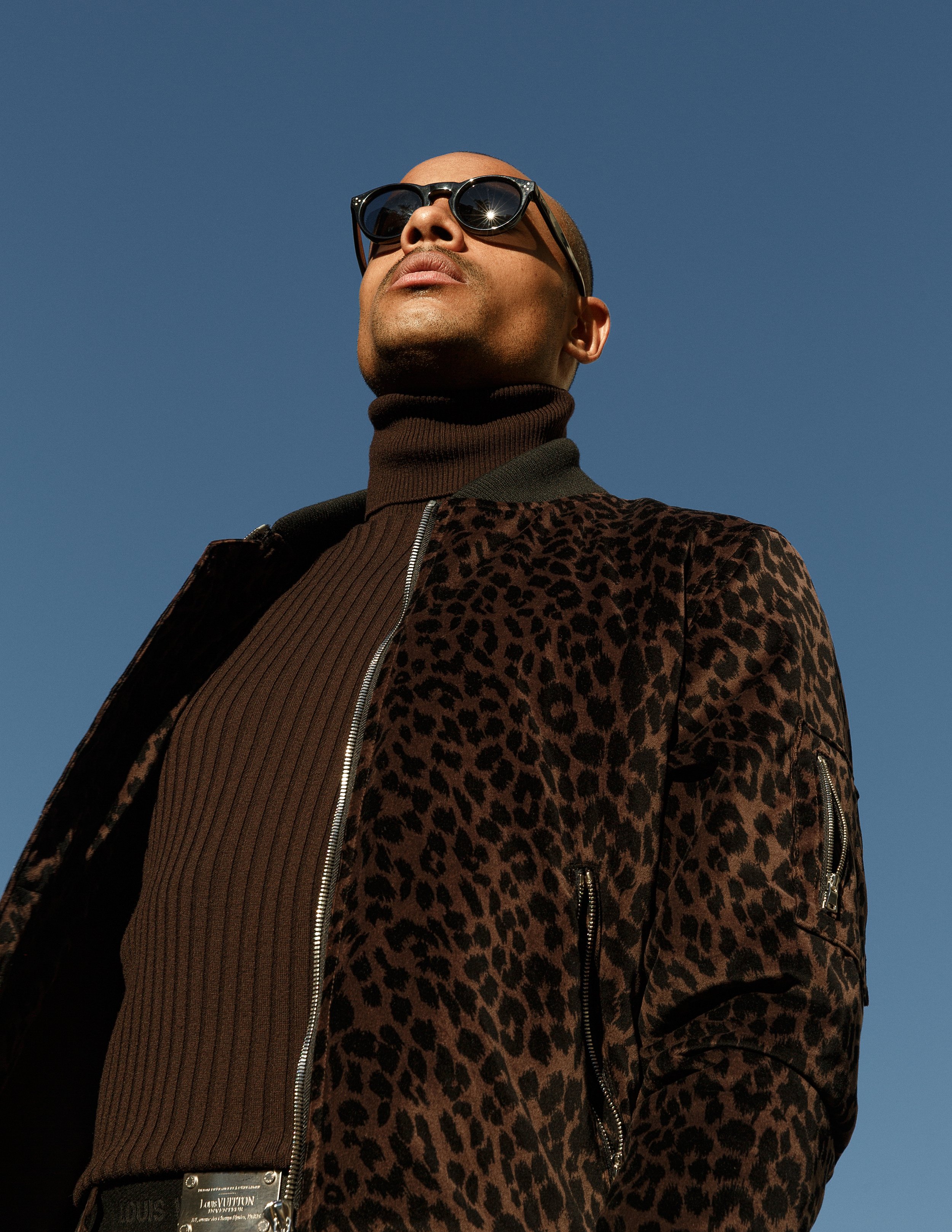 A person with a shaved head wearing dark sunglasses, a brown turtleneck shirt, and a leopard print jacket, against a clear blue sky.
