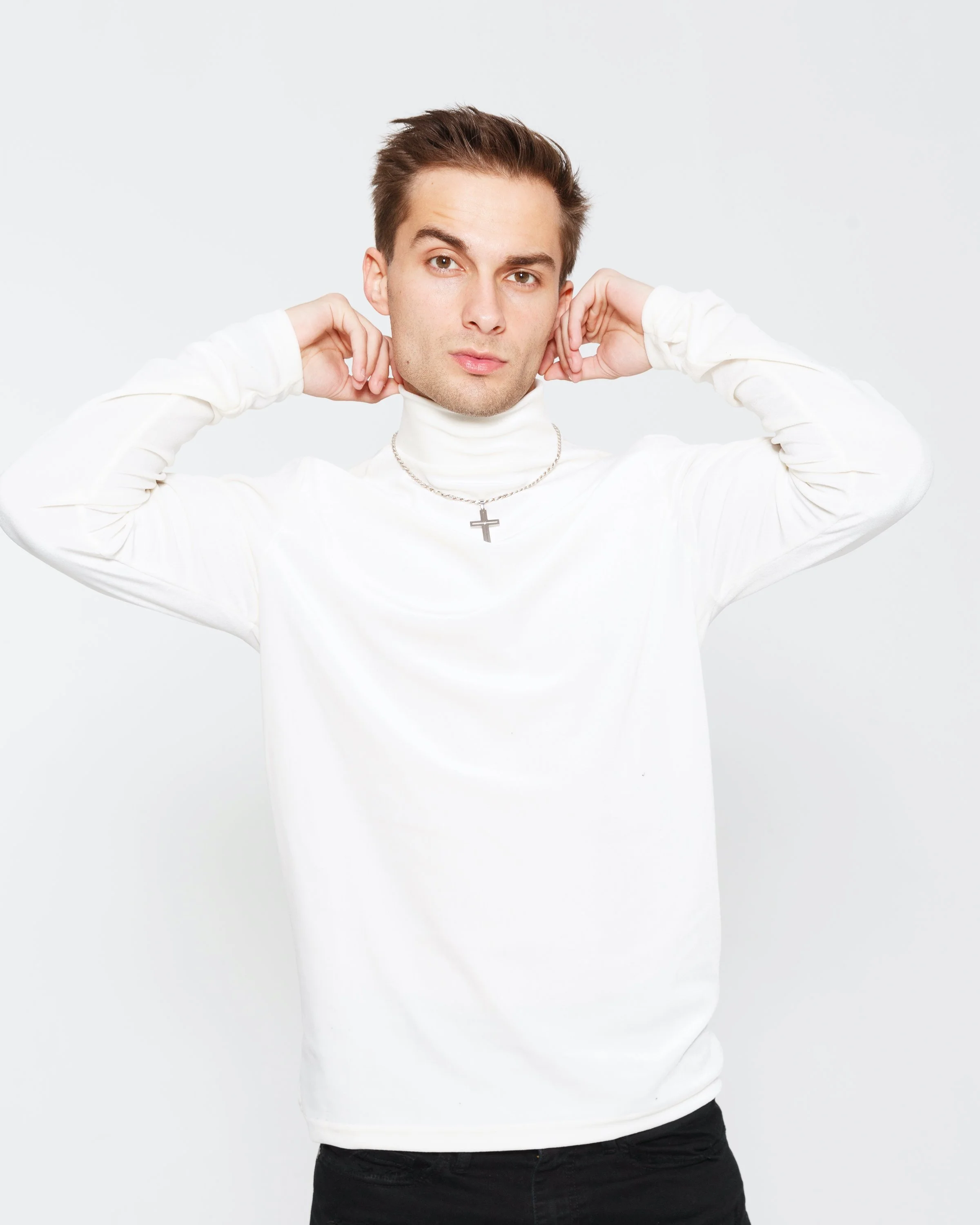 A young man with short brown hair and light skin, wearing a white turtleneck top and a silver cross necklace, is posing with his hands adjusting the collar of his shirt against a plain white background.