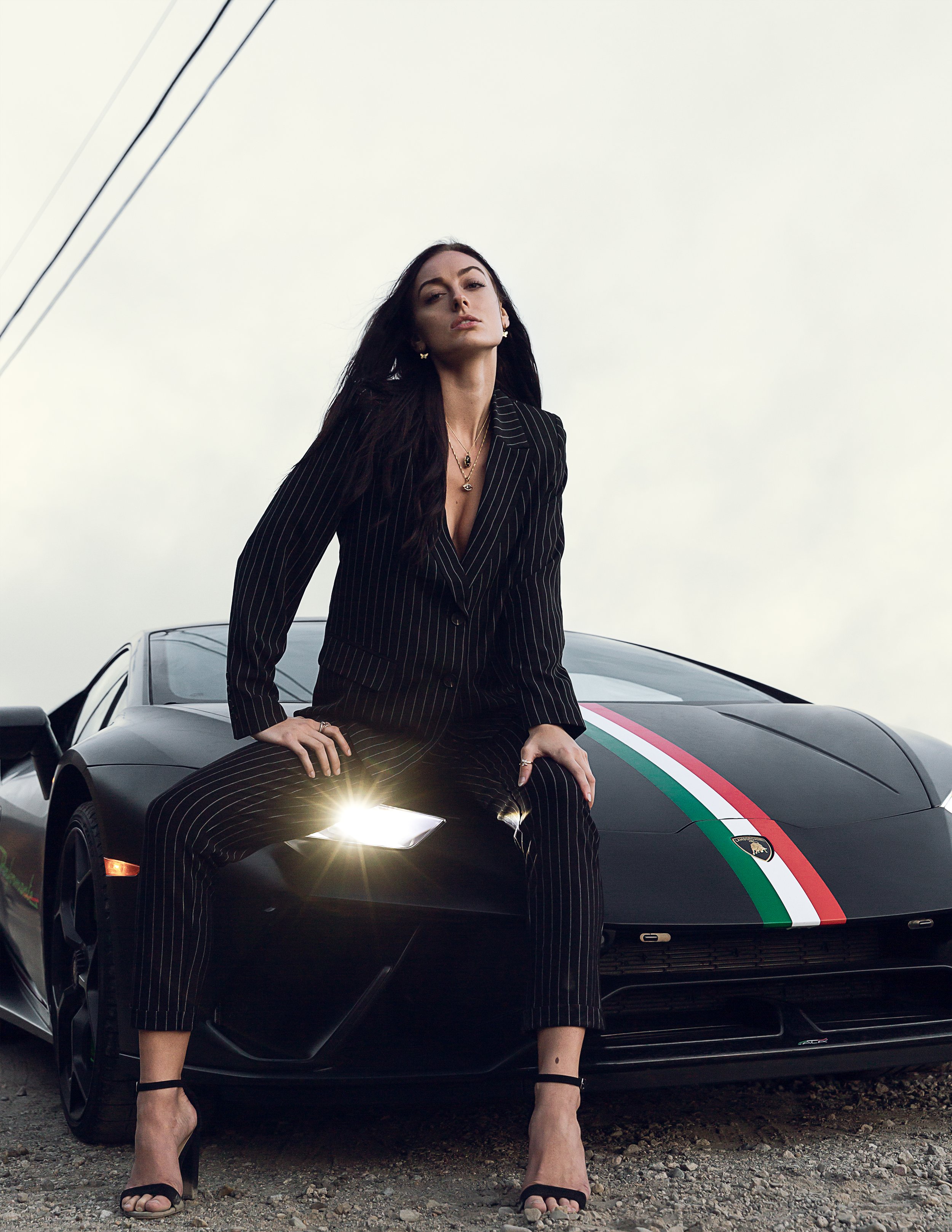 A woman in a black pinstripe suit with gold jewelry and high heels, sitting on the hood of a black sports car with Italian racing stripes, in an outdoor setting with gray overcast sky.