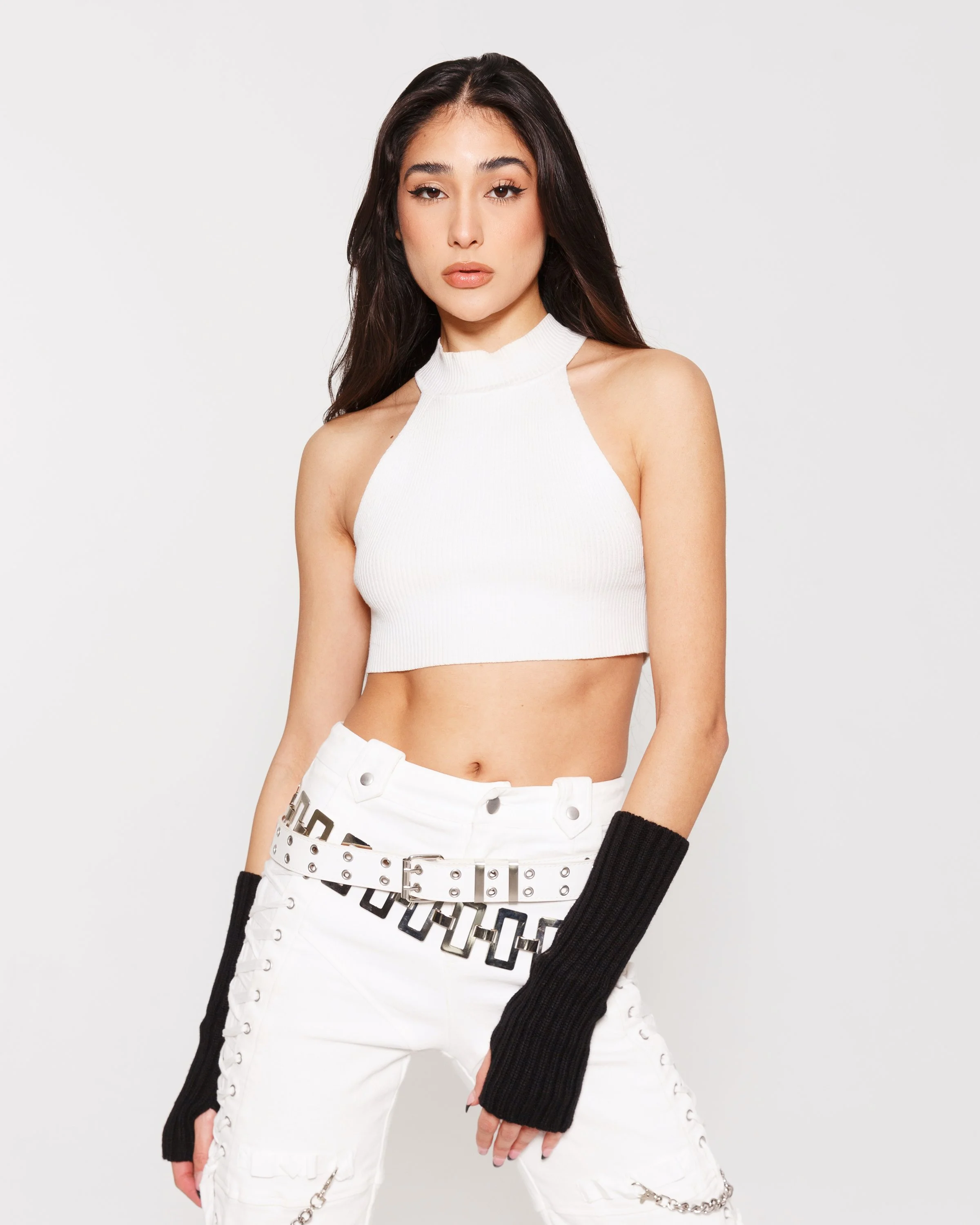 Young woman with long dark hair wearing a white sleeveless crop top, white pants with chain accents, black arm warmers, and posing against a light grey background.