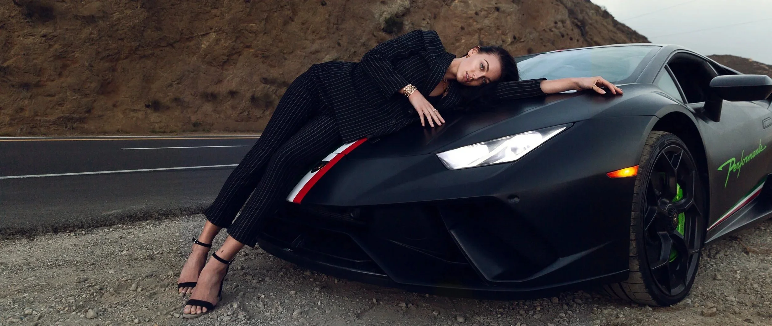 A woman in a black pinstripe suit wearing high heels, is lying on the hood of a black Lamborghini sports car, on the side of an empty road with a dirt hillside in the background.