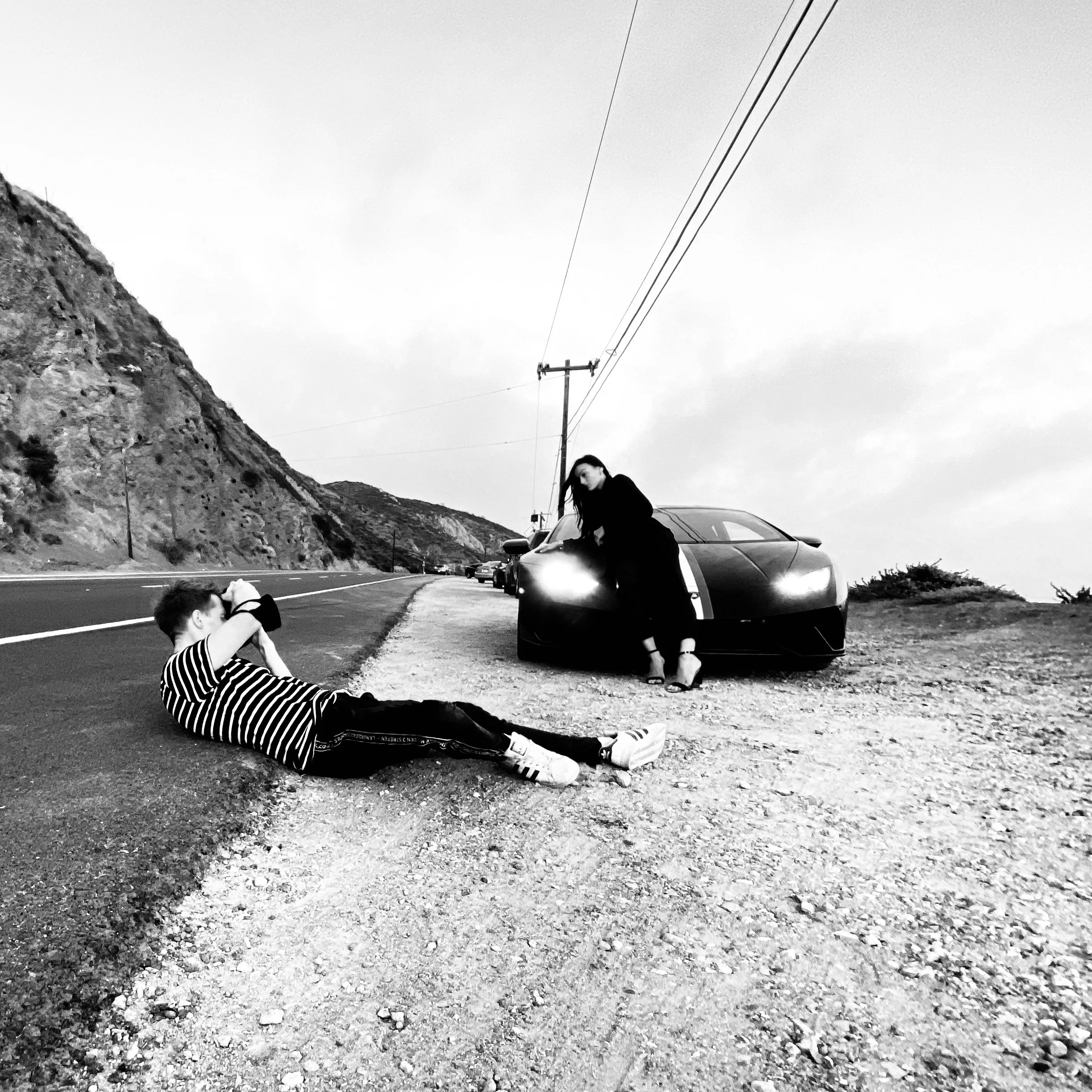 A person sitting on the ground near a parked car, with another person standing nearby, in a mountainous area under an overcast sky.