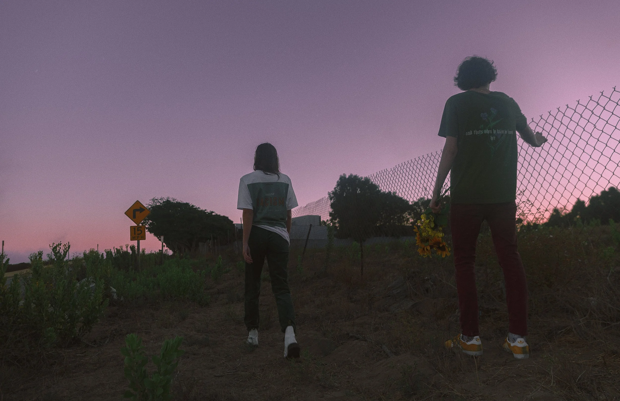 Two people walking away from the camera in a field at dusk, one holding flowers, with a chain-link fence on the right and trees in the background.