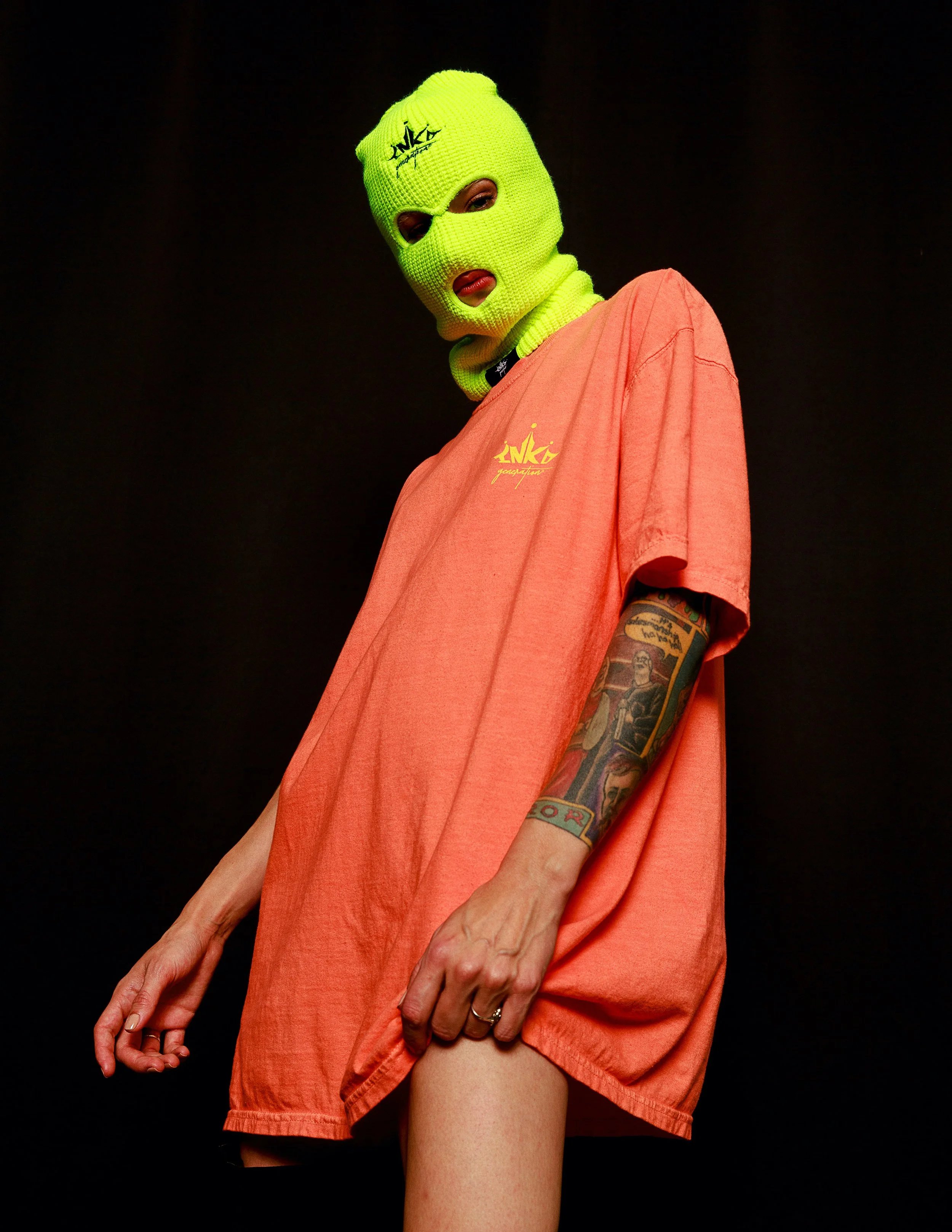 Person wearing a neon yellow ski mask and an oversized peach-colored t-shirt with tattoos visible on their arm, standing against a black background.