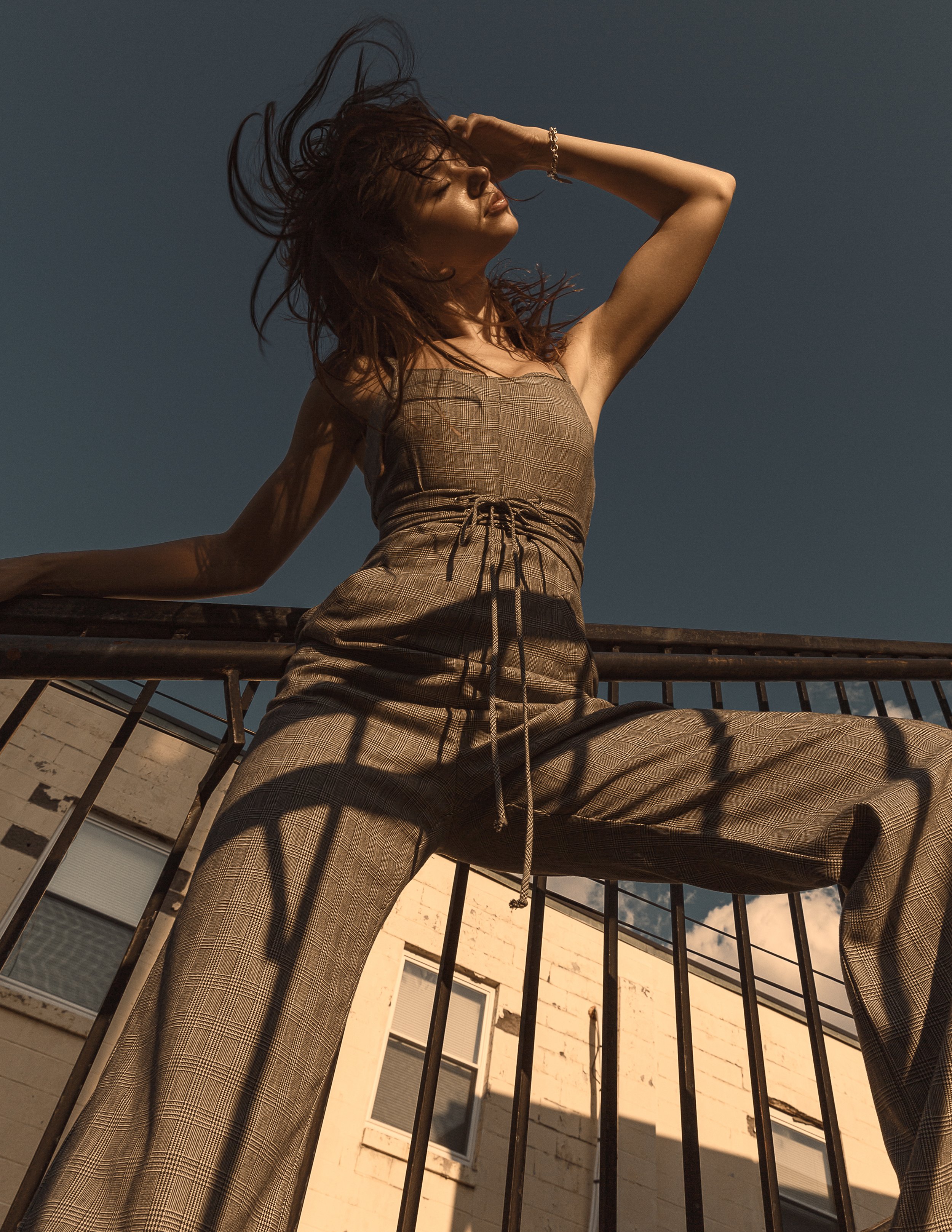 A woman with brown hair and a plaid outfit lifting her right arm, touching her head, with her eyes closed, standing behind a metal railing on a balcony. Sunlight casts shadows on her face. In the background, there is a building with windows and a partially visible sky.