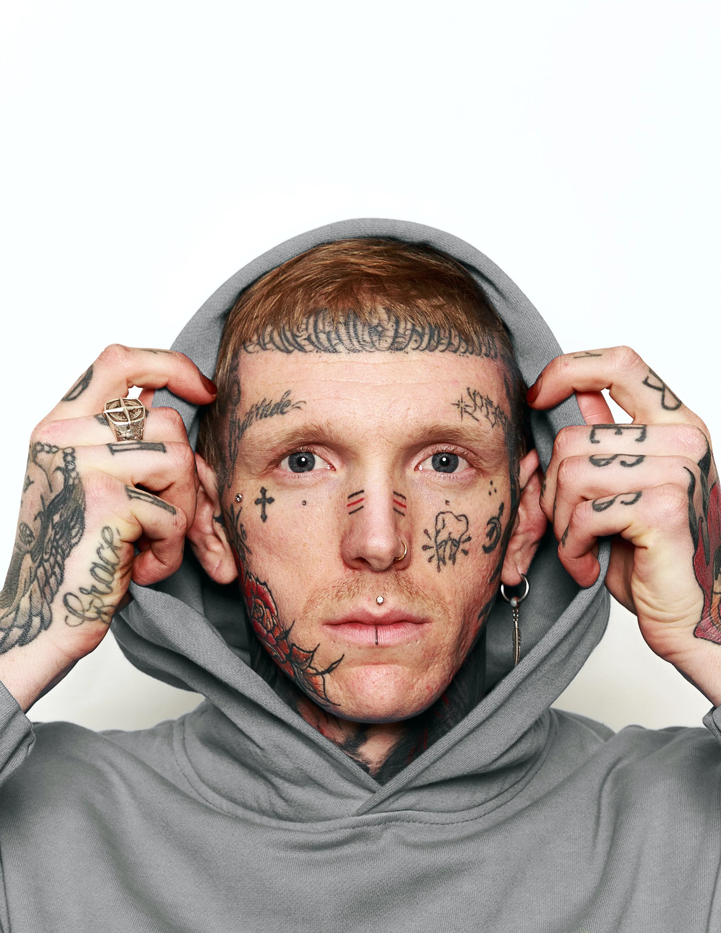 A person with short red hair and extensively tattooed face, hands, and neck, wearing a gray hoodie and holding the hood with both hands, with a white background.