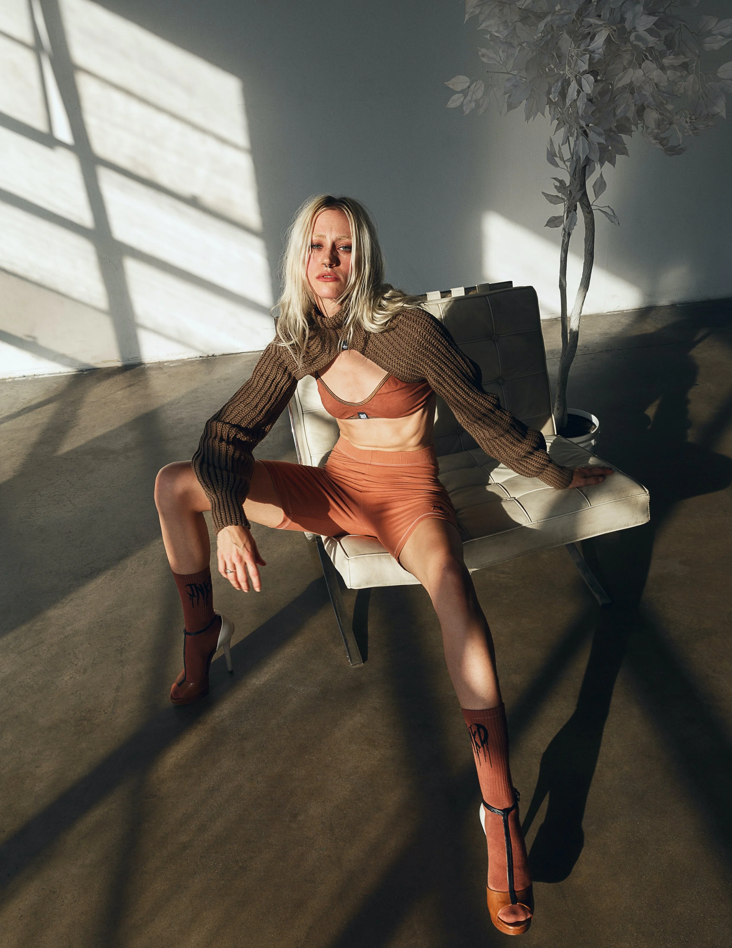 A woman with blonde hair wearing a brown mesh long-sleeve top, a matching brown bra, orange skirt, and orange socks with black text, posing on a beige chair in a room with sunlight casting shadows through a grid window.