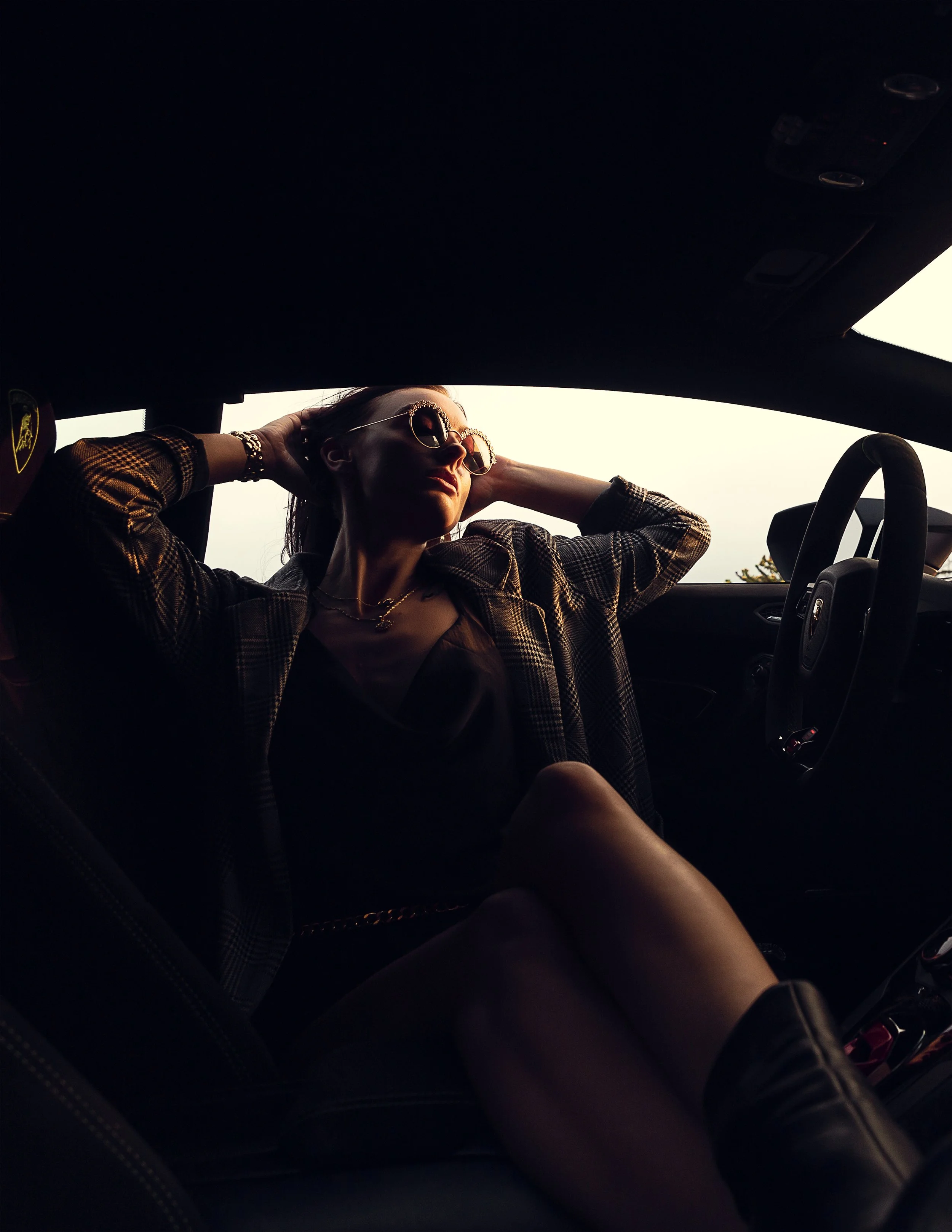 Woman sitting in a car seat with her hands behind her head, wearing sunglasses and a checkered jacket, looking out of the window.
