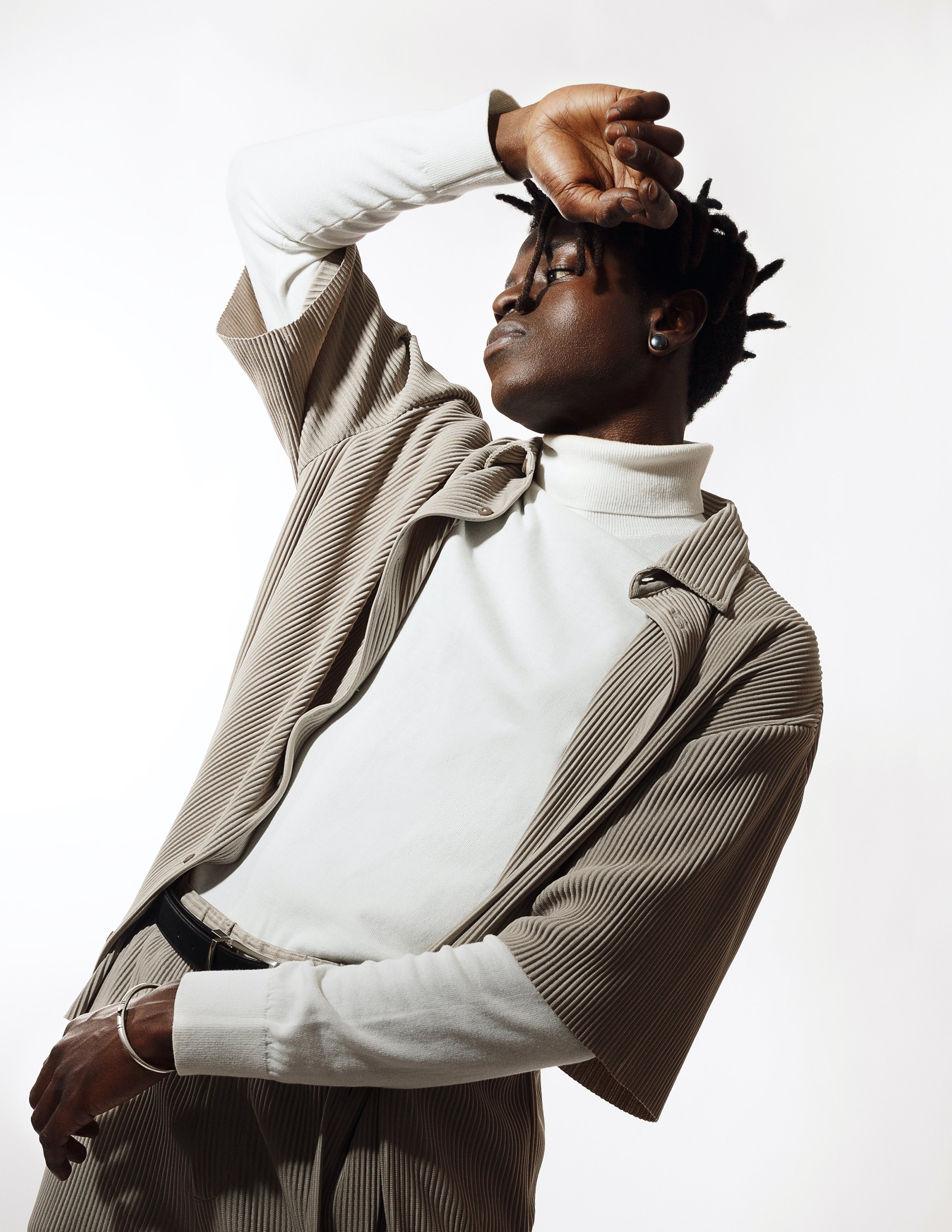 A person with dark skin and short dreadlocks wearing pearl earrings, a beige corduroy blazer, and a white turtleneck sweater, standing against a plain white background, with one arm raised above their head and the other resting on their hip.