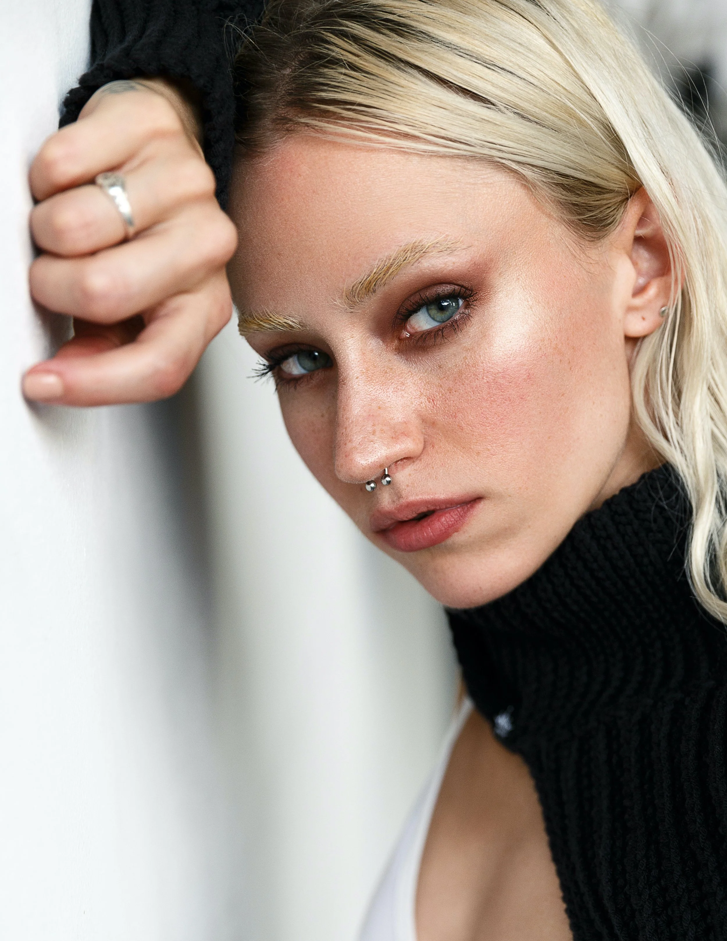 A close-up of a woman with blonde hair and piercing blue eyes, resting her head against a white wall, with her hand partially covering her face and showing a ring.