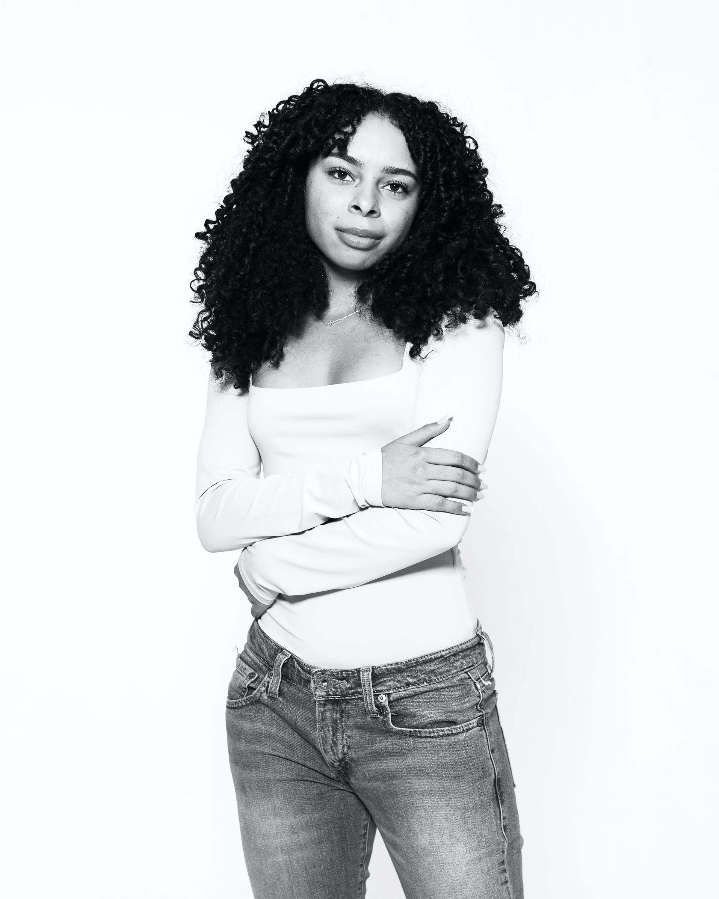 Black and white photograph of a woman with curly hair, wearing a white long sleeve shirt and jeans, posing with her arms crossed