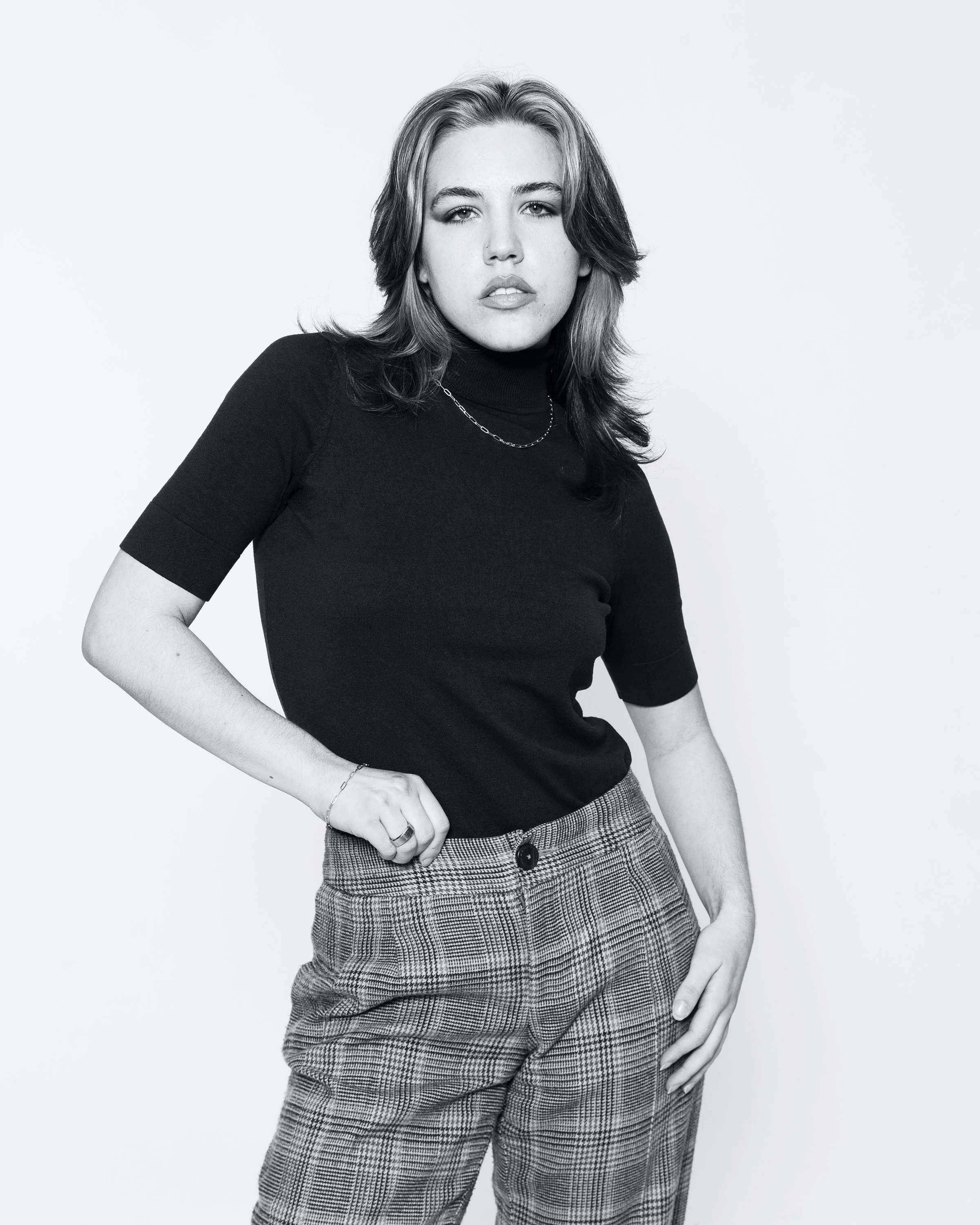 Black and white portrait of a woman with shoulder-length hair, wearing a black turtleneck and patterned pants, standing against a plain light background.
