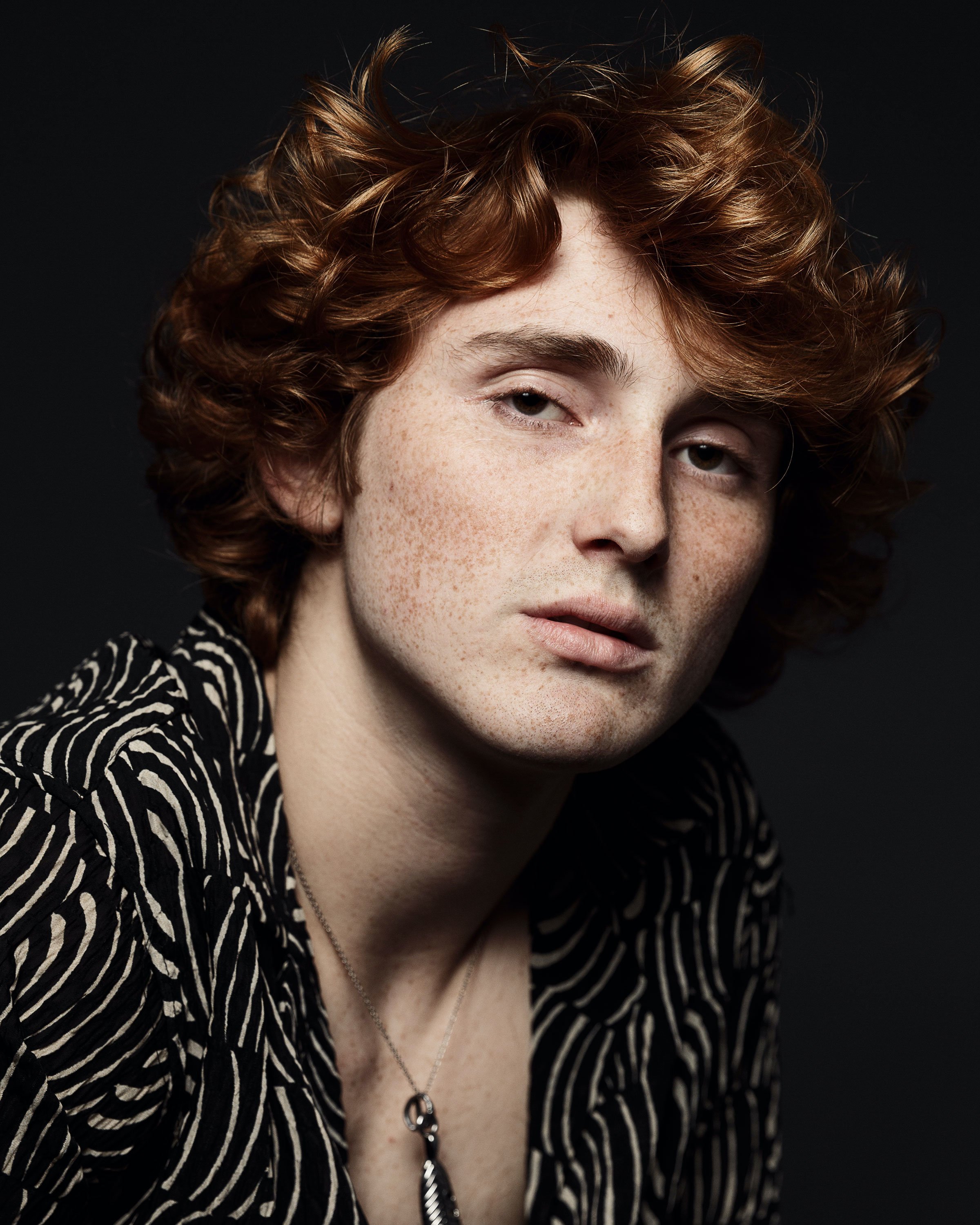 Close-up of a young woman with curly red hair, freckles, and light eyes, wearing a black and white striped shirt and a necklace with a pendant, against a dark background.