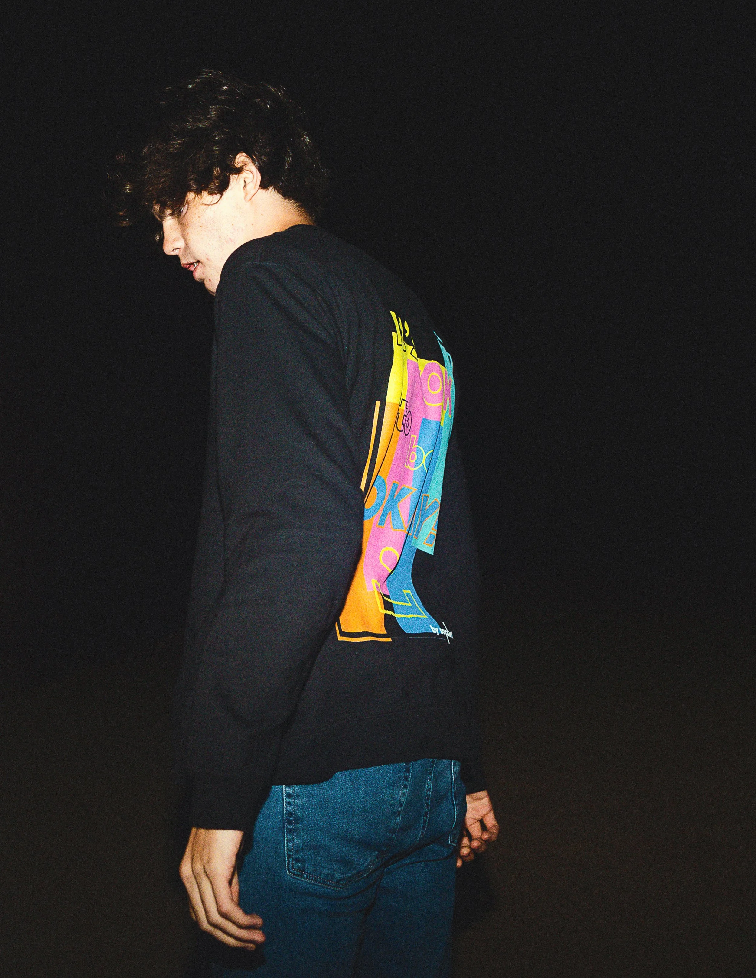 A person with curly dark hair wearing a black long sleeve shirt with colorful text and graphics on the back, and blue jeans, standing against a black background.