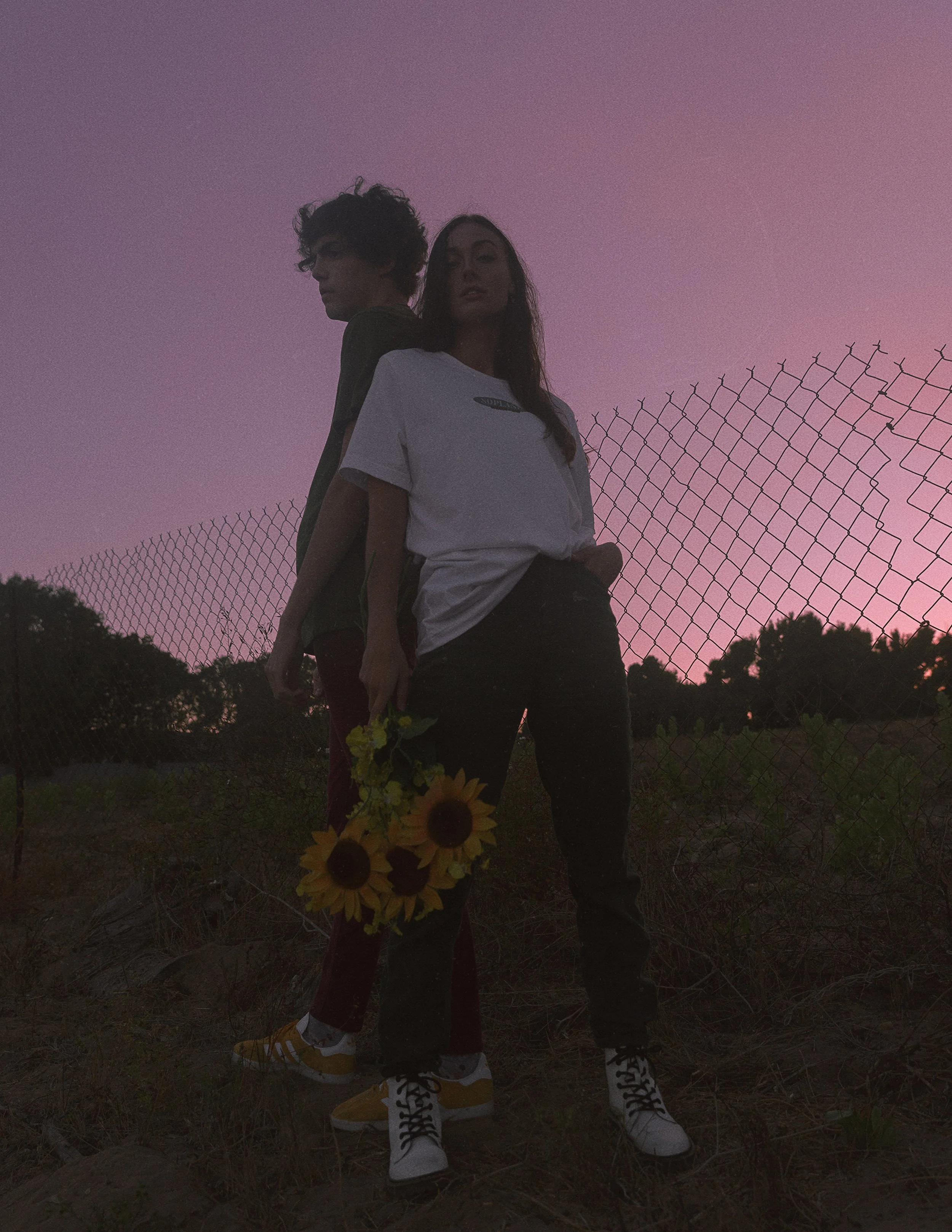 Two women standing back to back outdoors during sunset with a purple sky and a chain-link fence in the background. One woman is holding sunflowers.