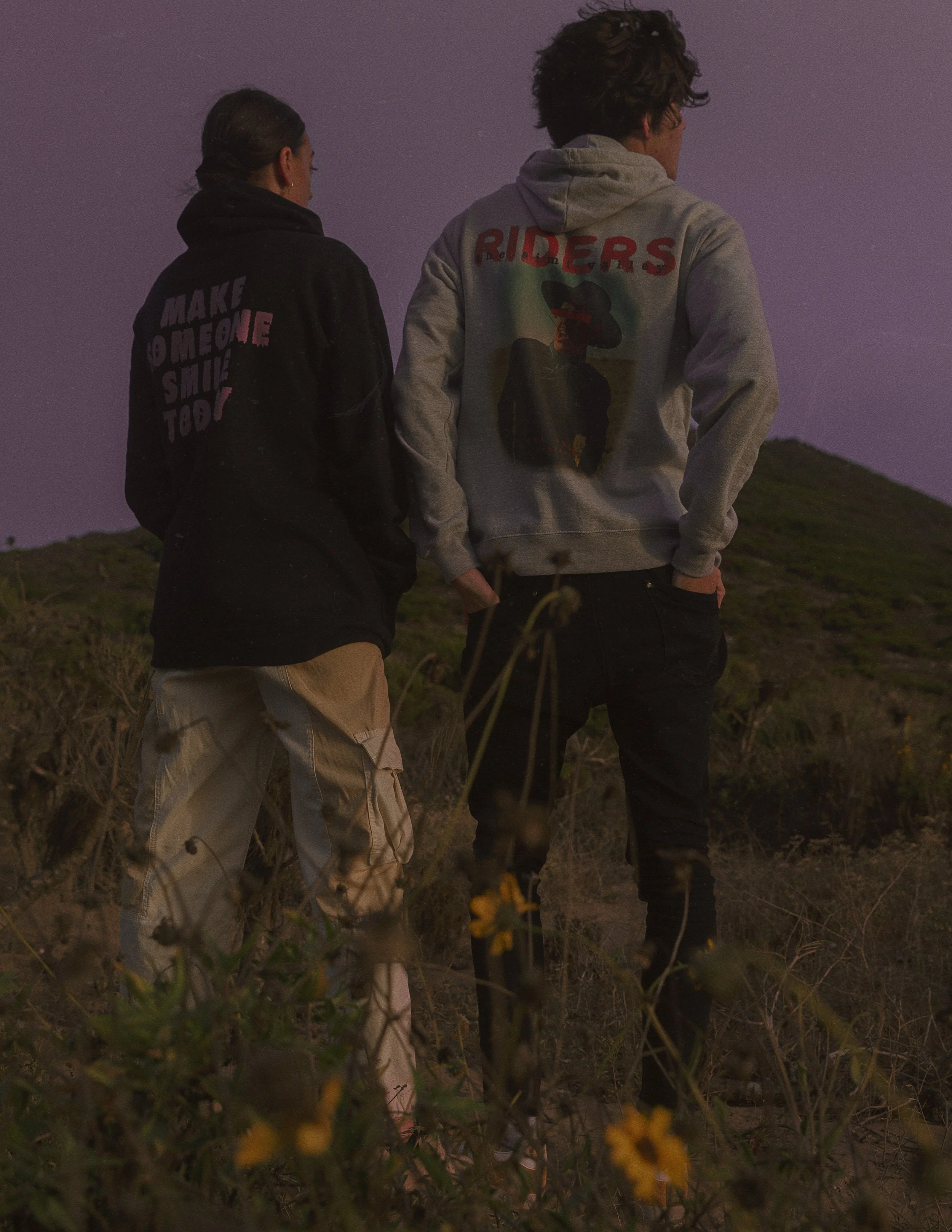 Two young people walking outdoors through grassy terrain during twilight, wearing hoodies with graphic designs and slogans, surrounded by yellow flowers and bushes.