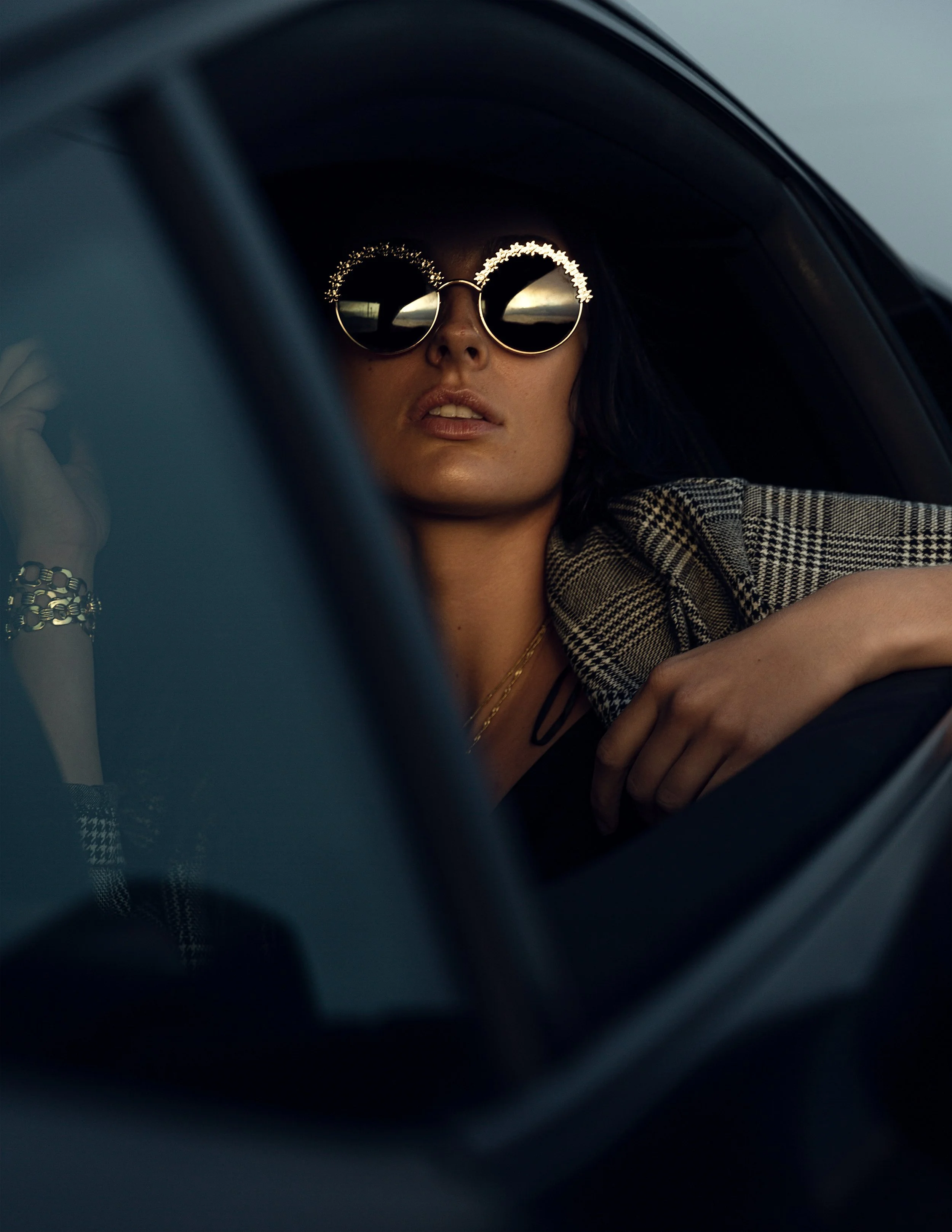A woman wearing sunglasses with floral details reflects a sunset, seated in a car, wearing a checkered blazer and gold jewelry.