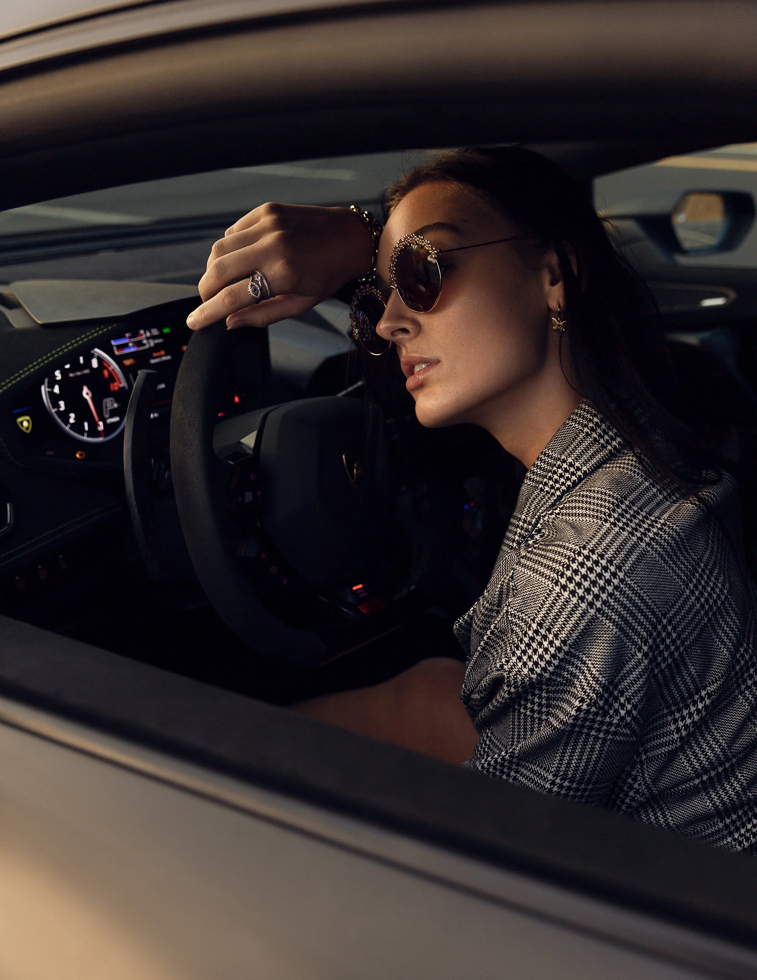 A woman wearing sunglasses and a checkered blazer is resting her head on her hand inside a car, with her eyes closed.