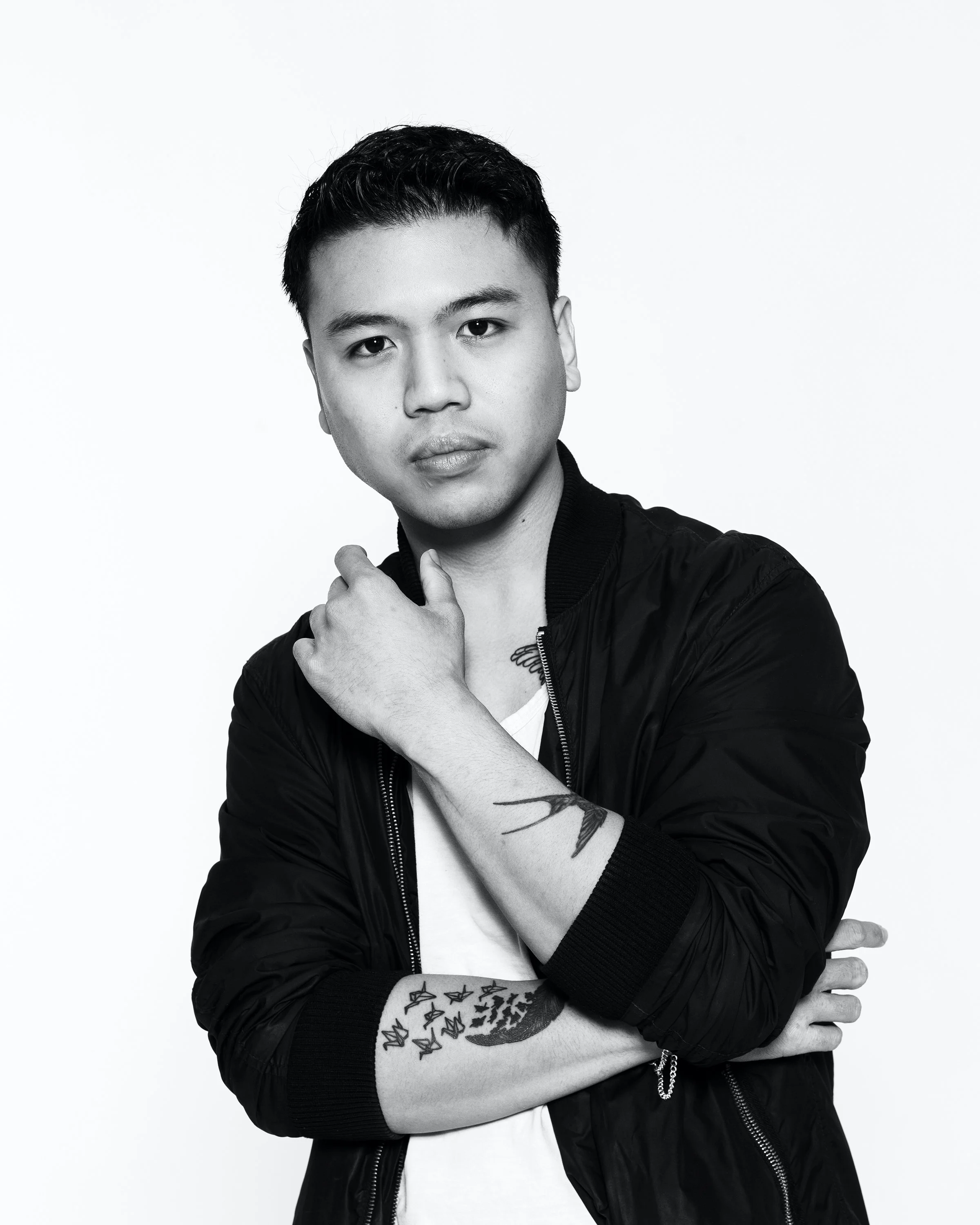 A young Asian man with short dark hair, looking serious, wearing a black jacket and white shirt, with tattoos on his arm, standing against a white background.