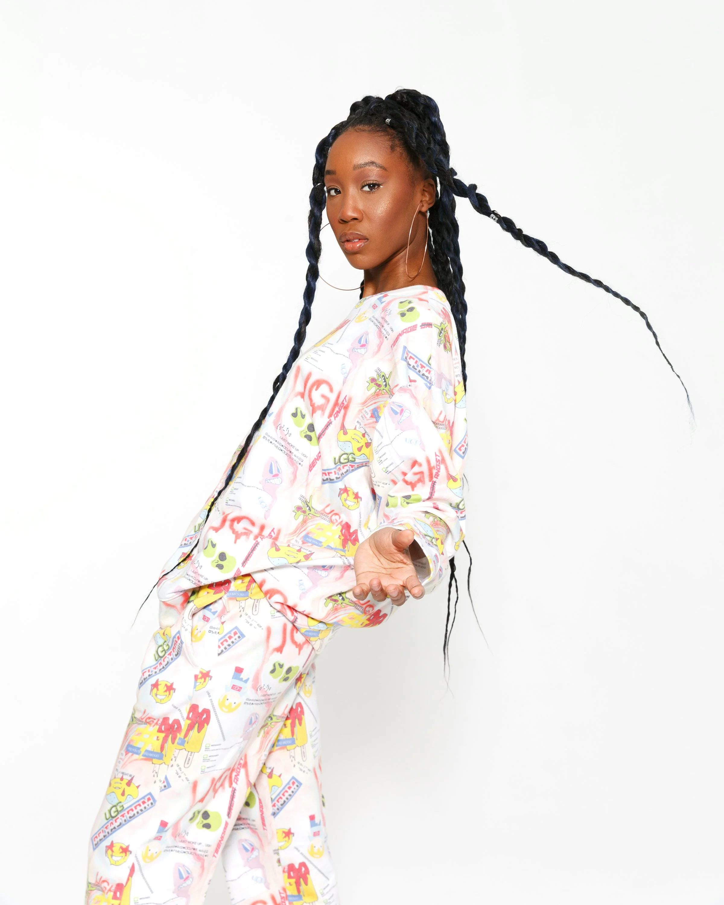 A woman with long braided black hair, wearing large hoop earrings, posing against a plain white background. She is dressed in a loose-fitting, colorful, patterned garment featuring cartoon characters and various graphic designs. She is looking at the camera with a neutral expression, her left hand extended outward.