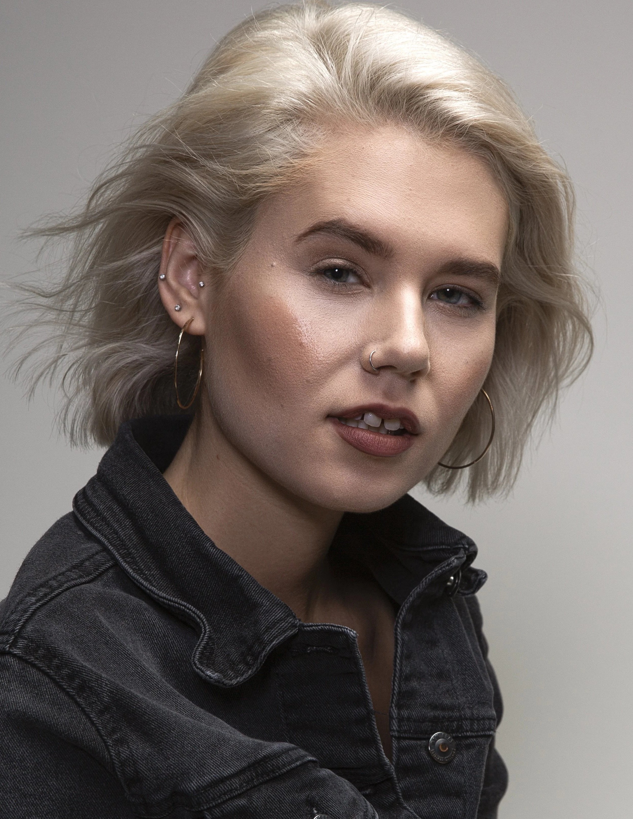 A close-up of a woman's face with platinum blonde hair, wearing a black denim jacket, hoop earrings, and a nose ring, against a gray background.