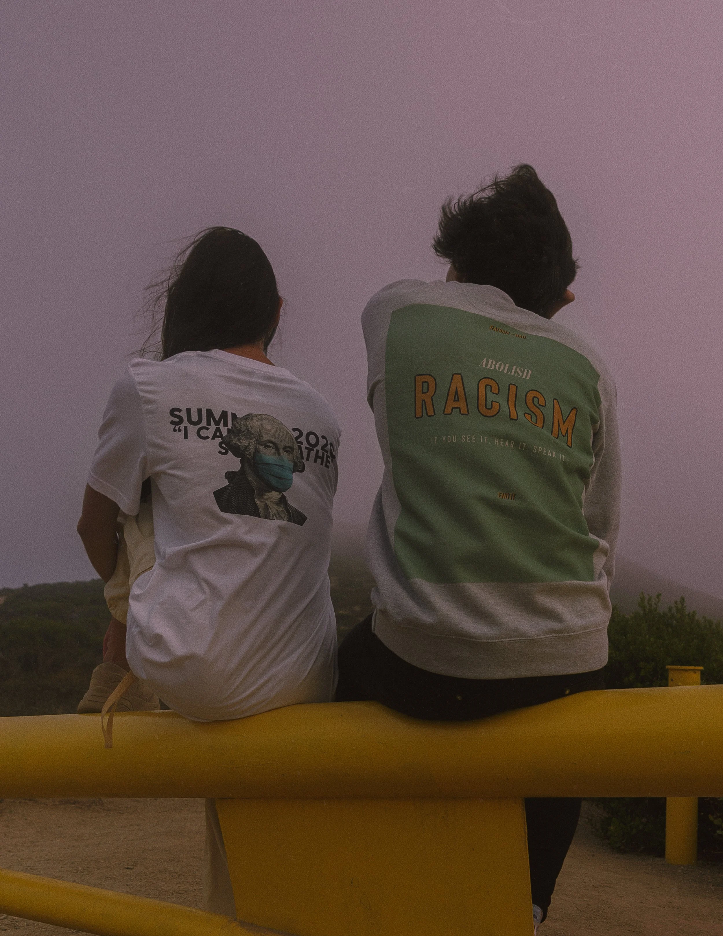 Two people sitting on a yellow boat, facing away, at twilight. One person wears a white t-shirt with image of a woman wearing a face mask, and the other person wears a green t-shirt with the word 'RACISM' and the phrase 'ABOLISH RACISM' on the back.