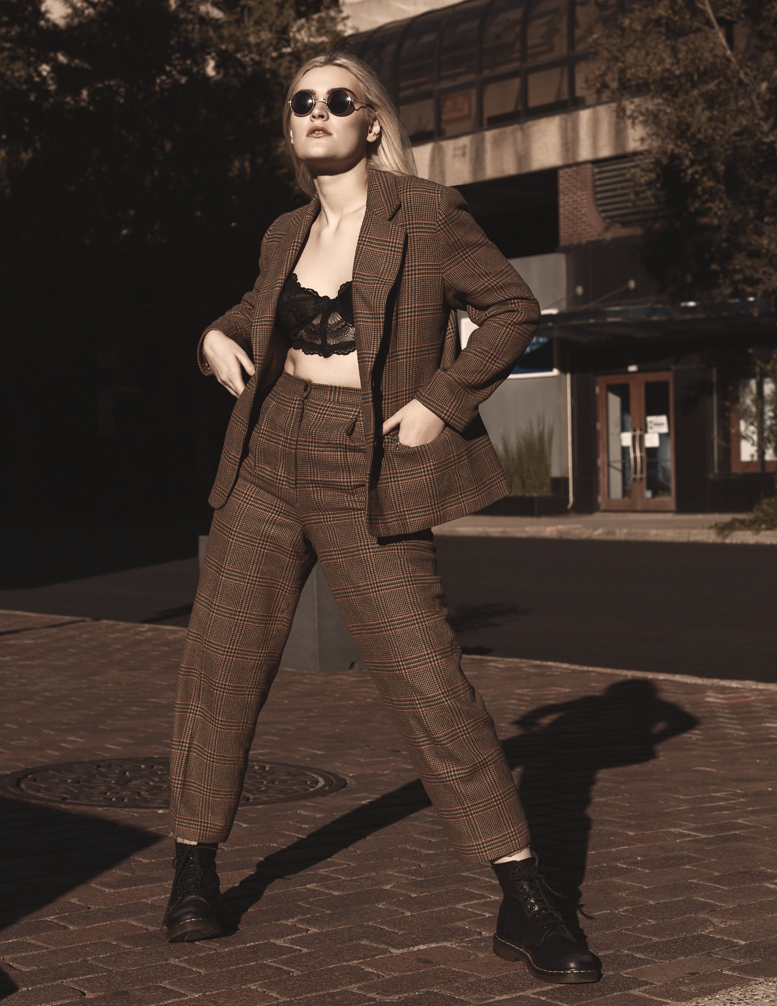 A woman standing outdoors at night wearing sunglasses, a brown plaid suit with a black lace top, and black combat boots, striking a confident pose.