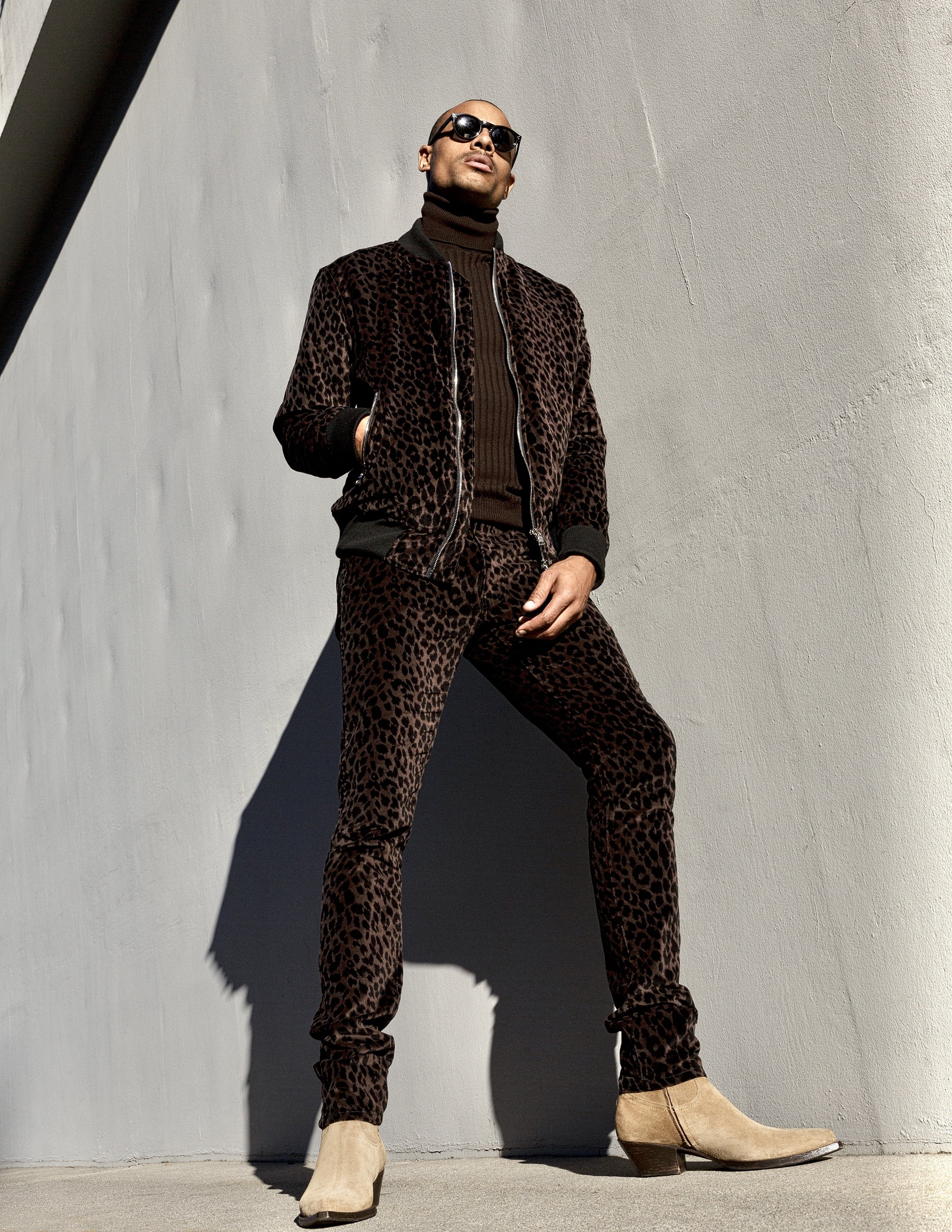 A man wearing a leopard print jacket and matching pants, with beige cowboy boots, stands against a plain wall, looking upwards with his hands in his coat pockets, wearing sunglasses.