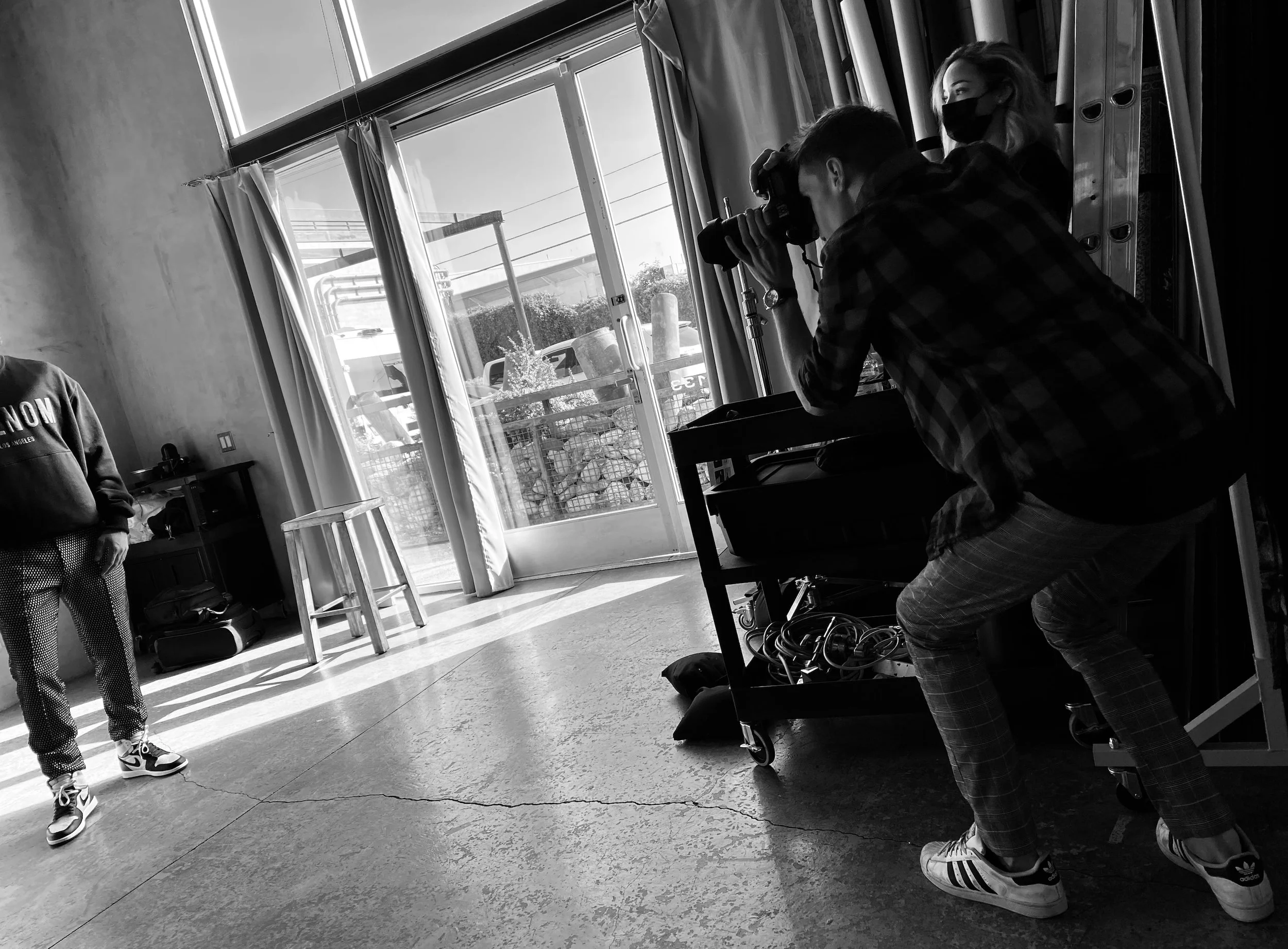 A person wearing a plaid shirt and sneakers is kneeling in front of a camera on a tripod, taking a photo inside a room with large windows and curtains. Another person, wearing a mask and casual clothes, is sitting on a bench nearby, looking to the right. The room has a polished floor and a door with glass panels that lets in natural light.