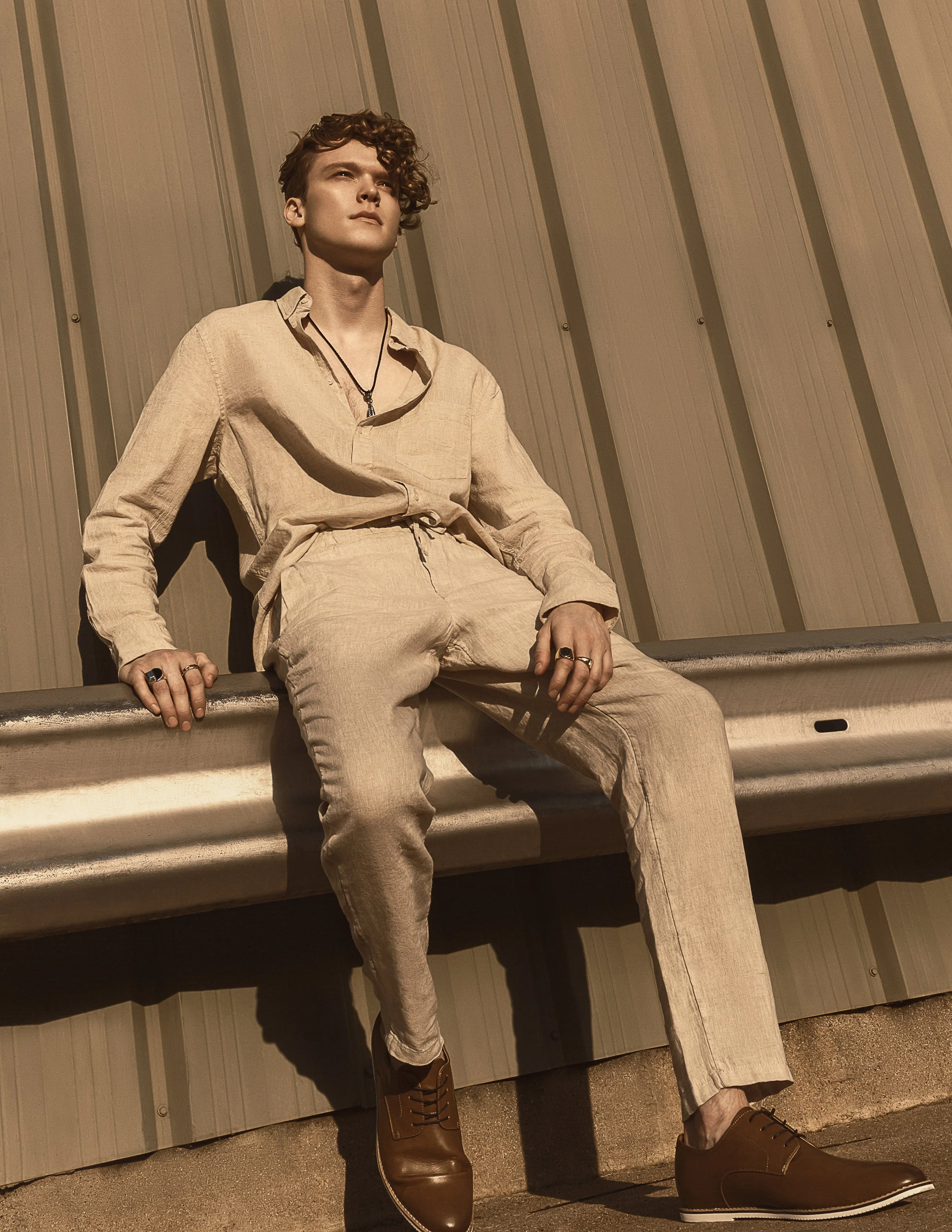 Young man in beige outfit sitting against a metal wall, wearing brown leather boots and rings on his fingers.