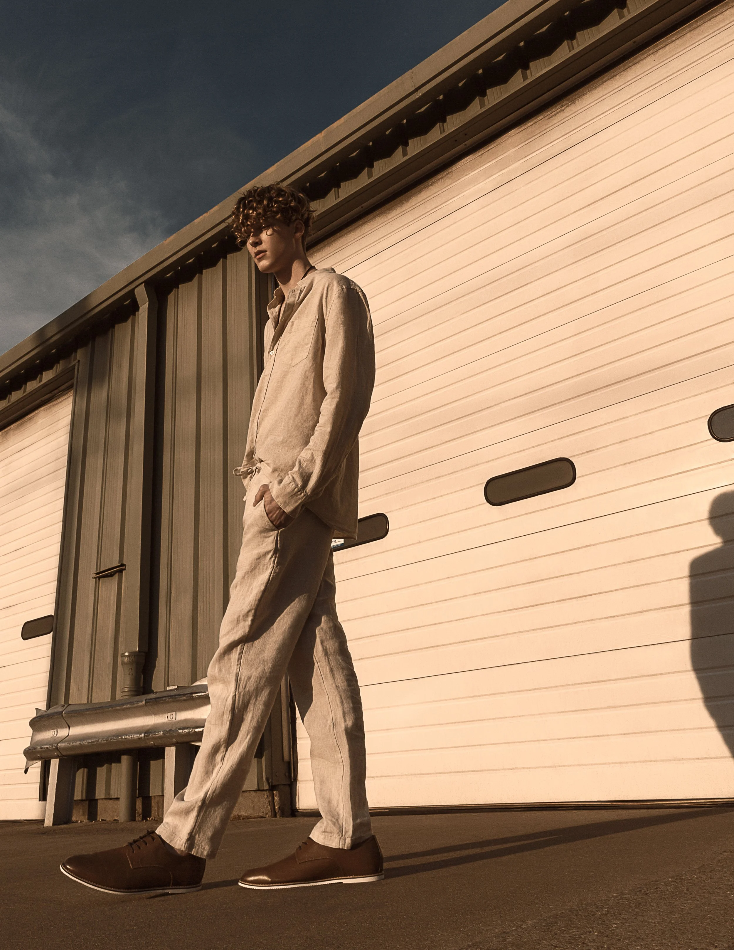 A young man with curly hair dressed in beige clothing and brown shoes standing outdoors near a beige garage door during sunset.