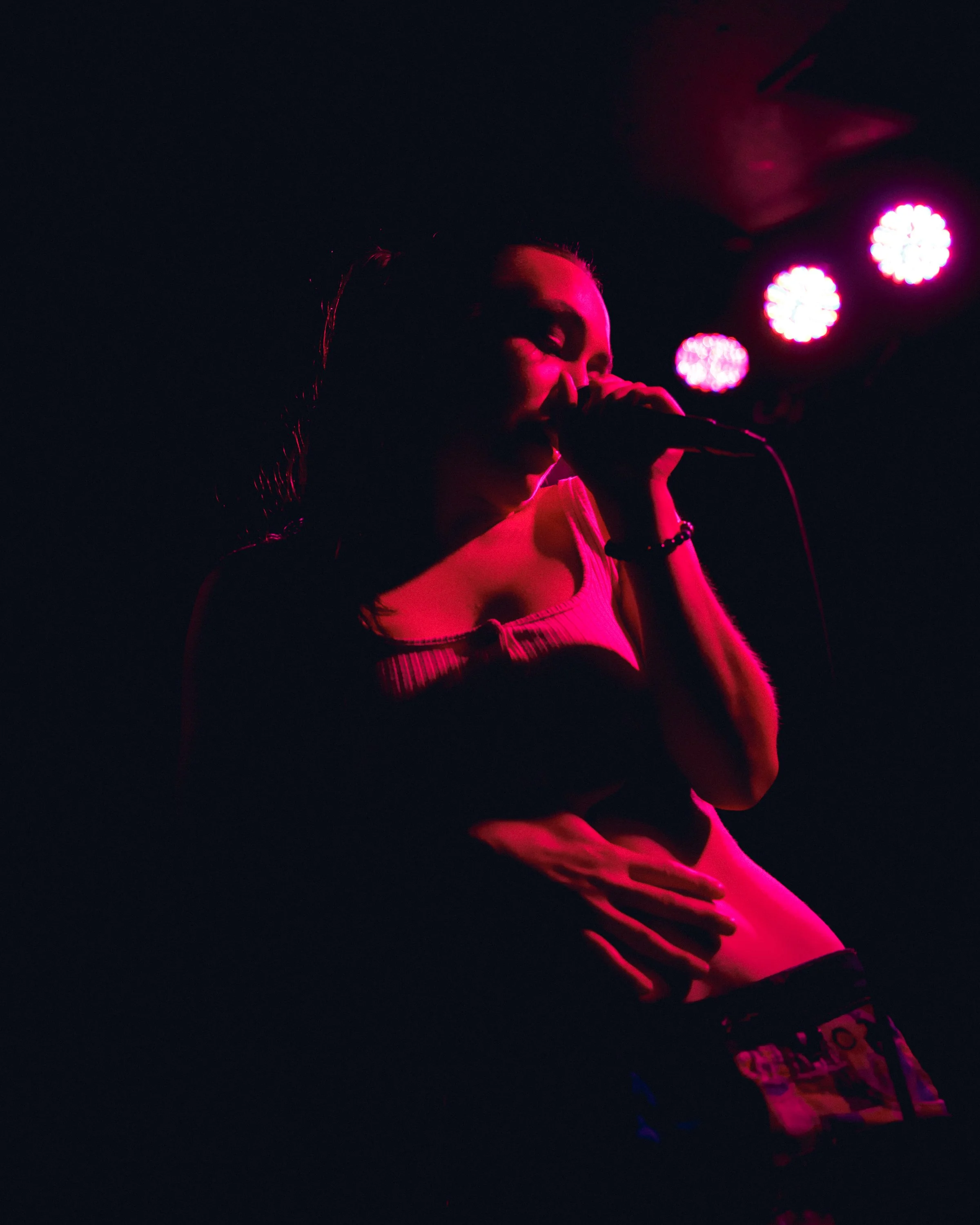 A woman singing into a microphone on stage illuminated by pink stage lights, with her hand resting on her stomach.