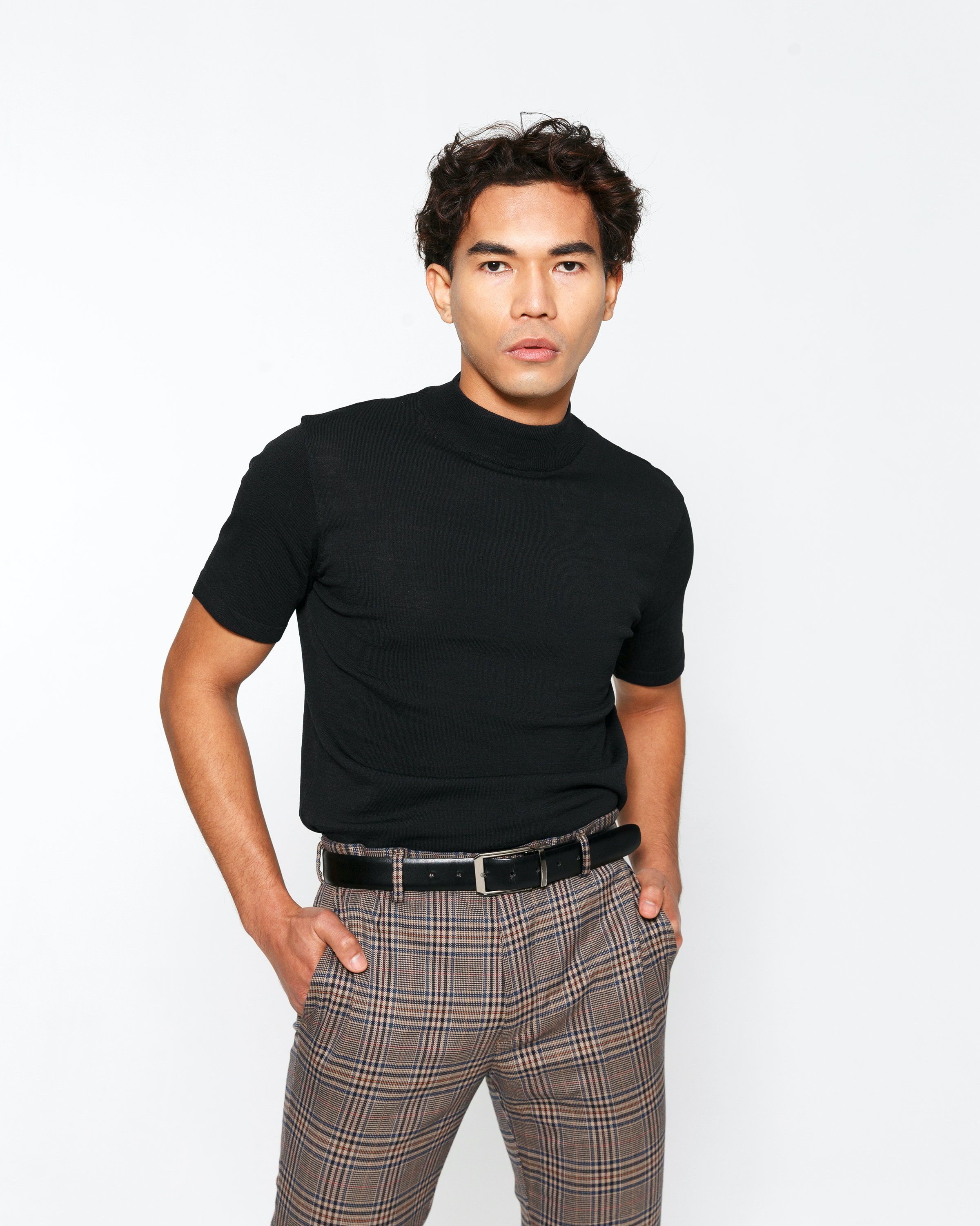 Young man in black turtleneck and plaid pants posing with hand in pocket against white background.