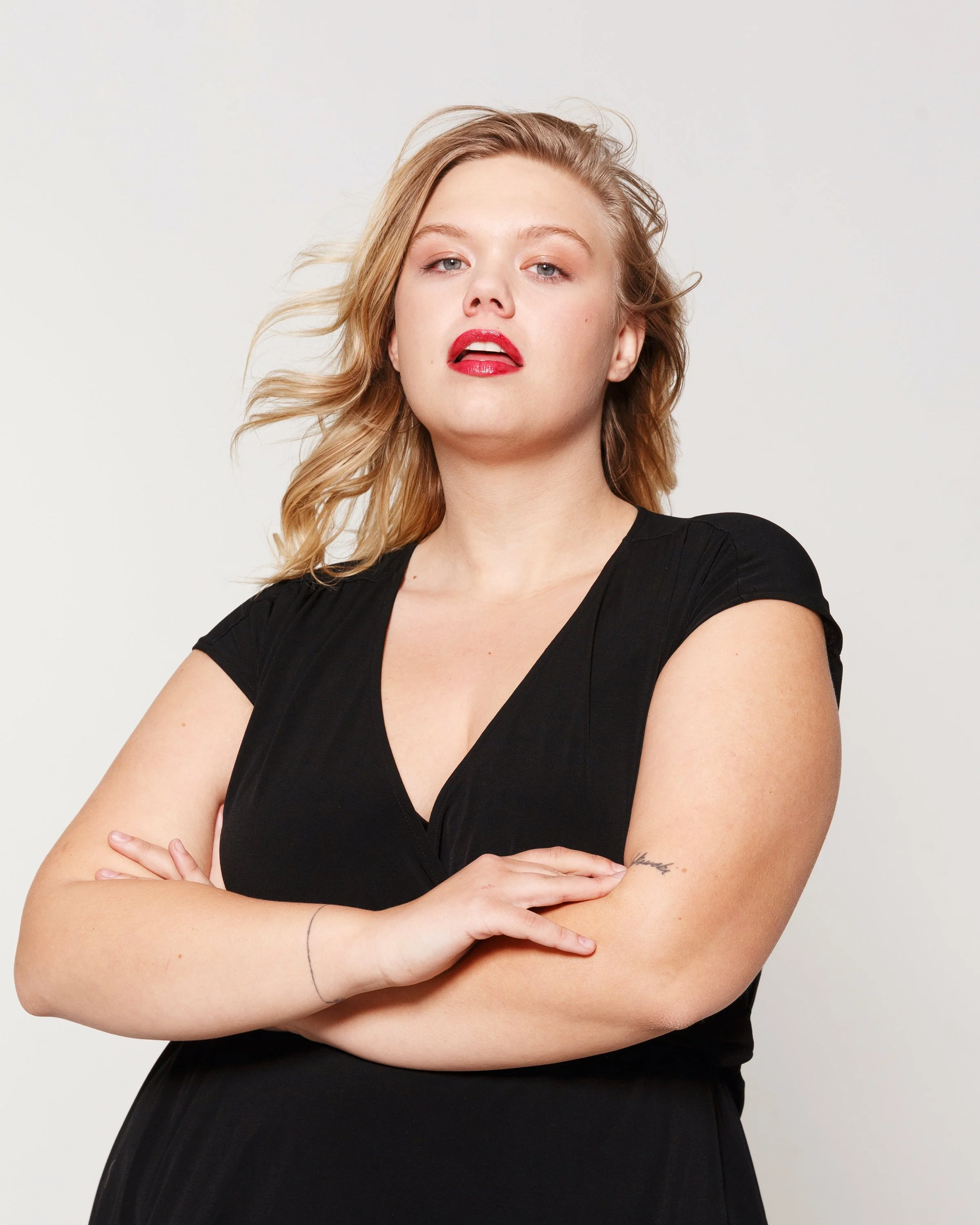 A woman with long blonde hair, wearing a black top, has her arms crossed and is looking confidently at the camera with red lipstick and natural makeup, against a plain light background.