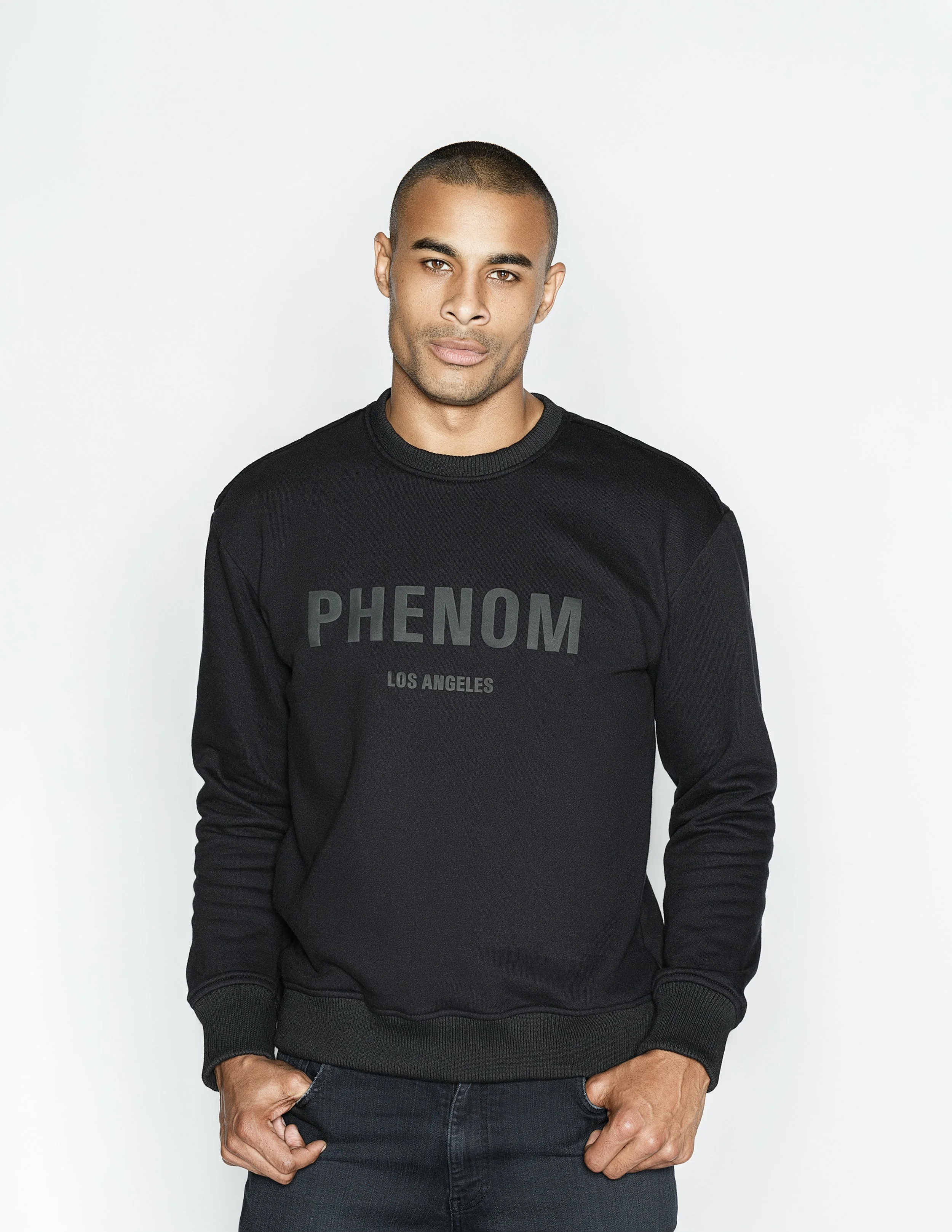 A man with tan skin and short black hair poses against a plain white background, wearing a black sweatshirt with the word 'PHENOM' and 'LOS ANGELES' written on it.