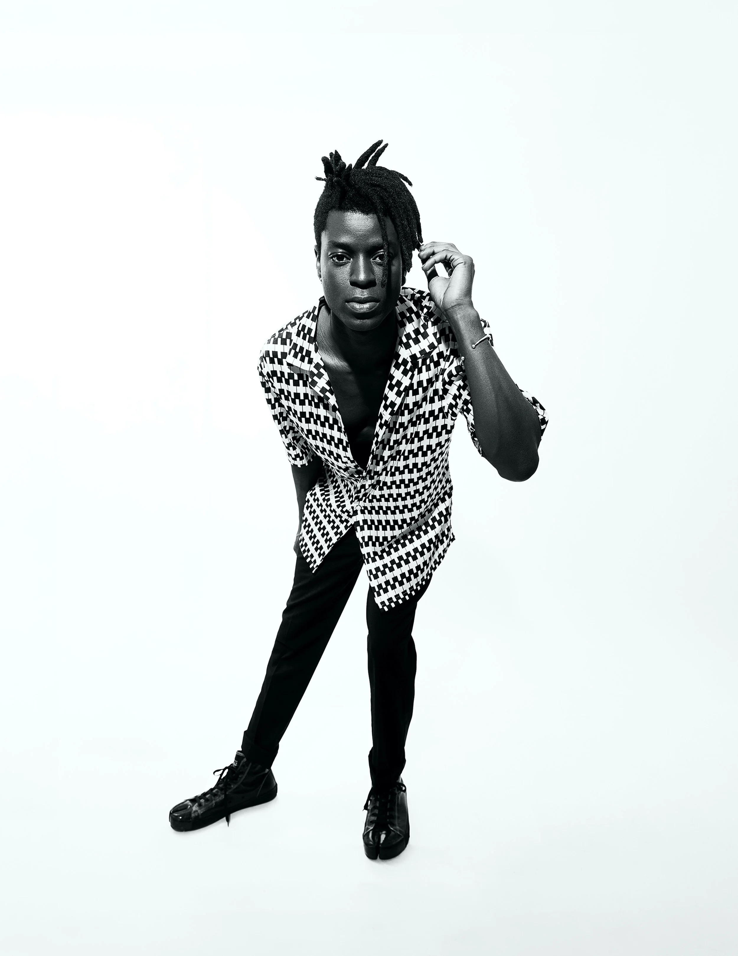A woman with dark skin and dreadlocks dressed in a patterned shirt, black pants, and black boots, leaning forward slightly and touching her hair with her right hand against a plain white background.