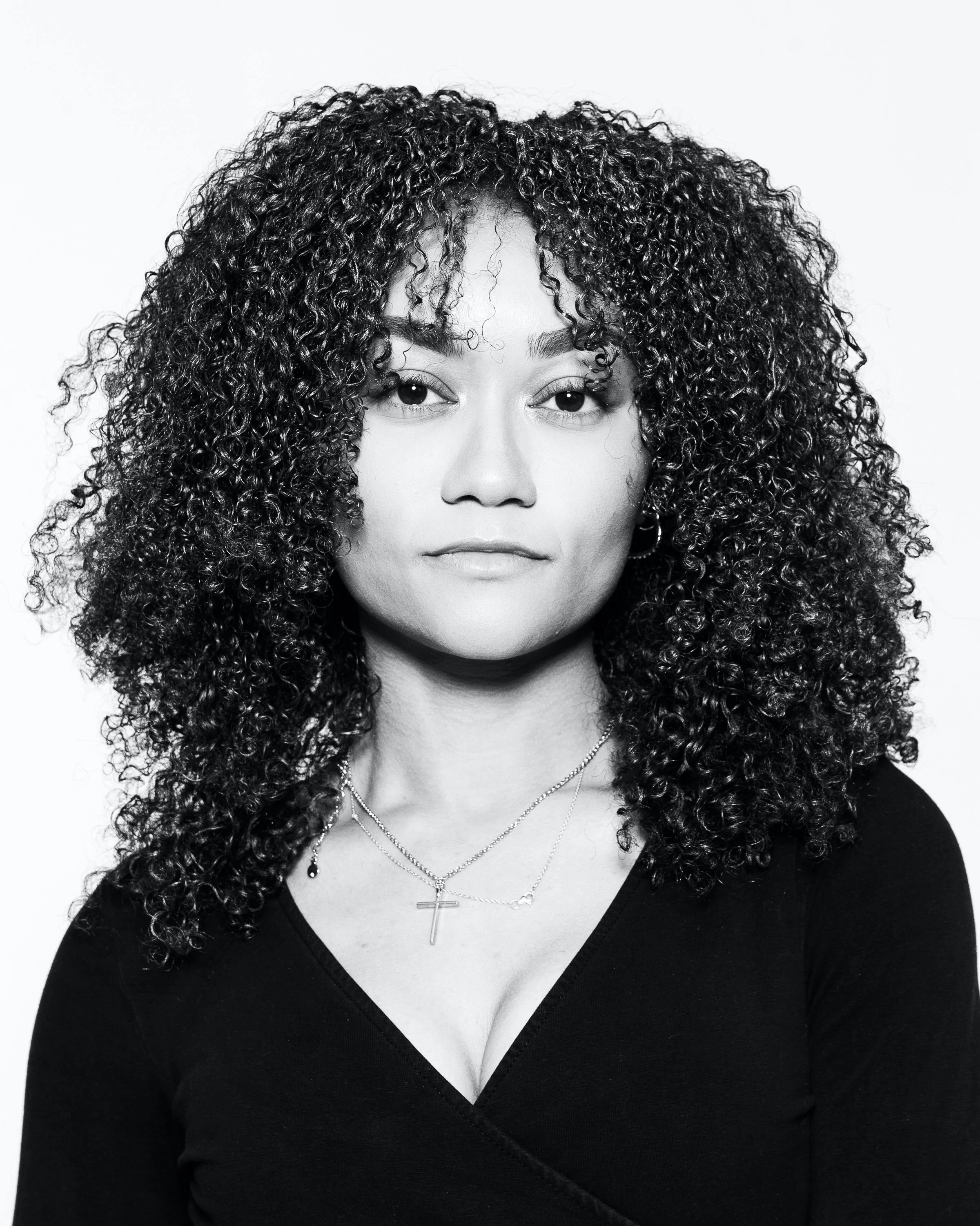 A woman with curly hair wearing a black top and layered necklaces, including a cross, against a plain background.