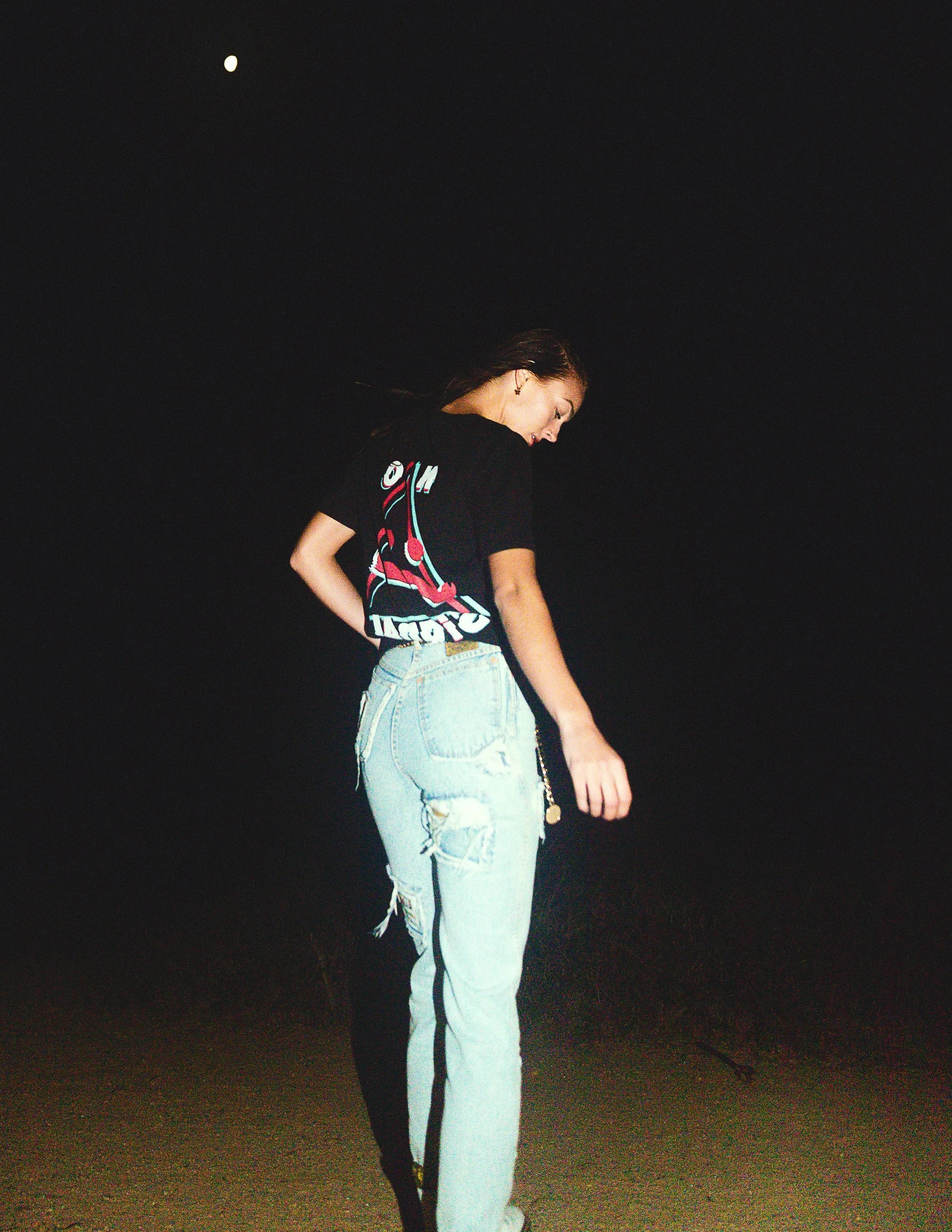 A young woman with long hair wearing a black T-shirt with colorful graphics and light blue distressed jeans standing outdoors at night.