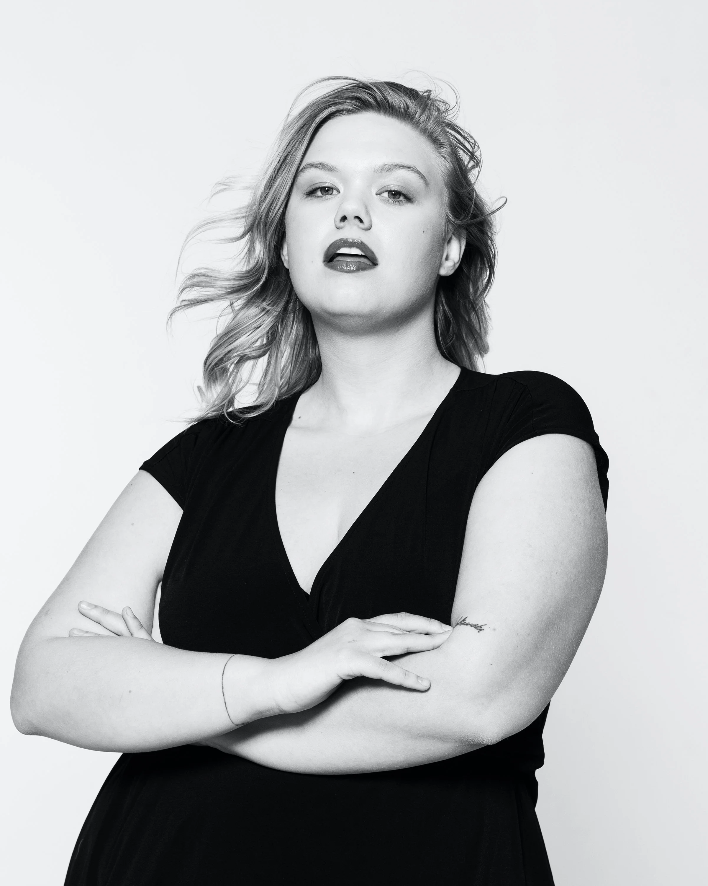 Black and white portrait of a woman with wavy hair, wearing a black top, with crossed arms, looking confidently into the camera.