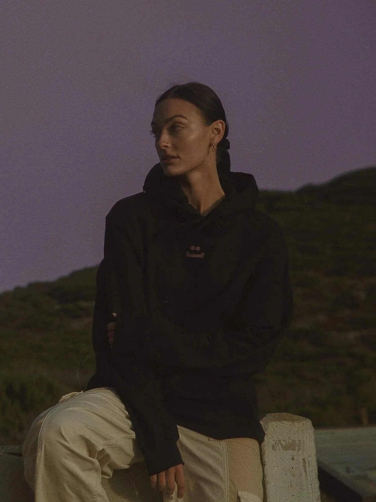 A woman with dark hair pulled back, wearing a black jacket and beige pants, sitting on a low concrete barrier outdoors during dusk or night, with hills and a purple sky in the background.