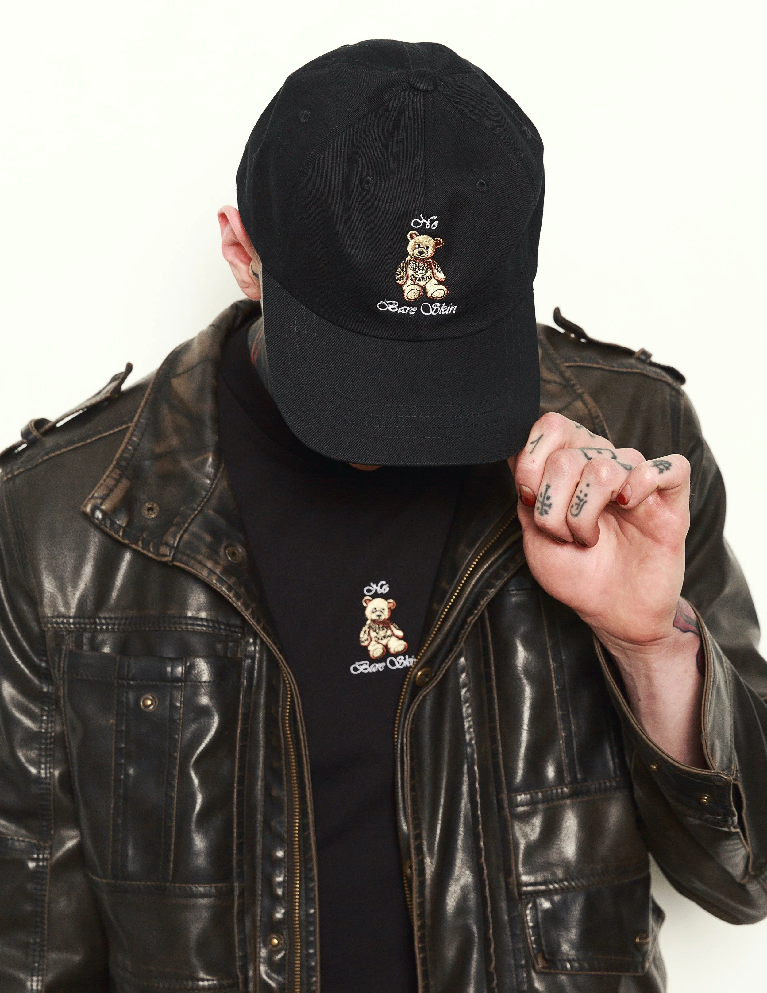 Person wearing a black baseball cap with an embroidered bear patch and the words 'Beare Oskin' on it, dressed in a brown leather jacket.
