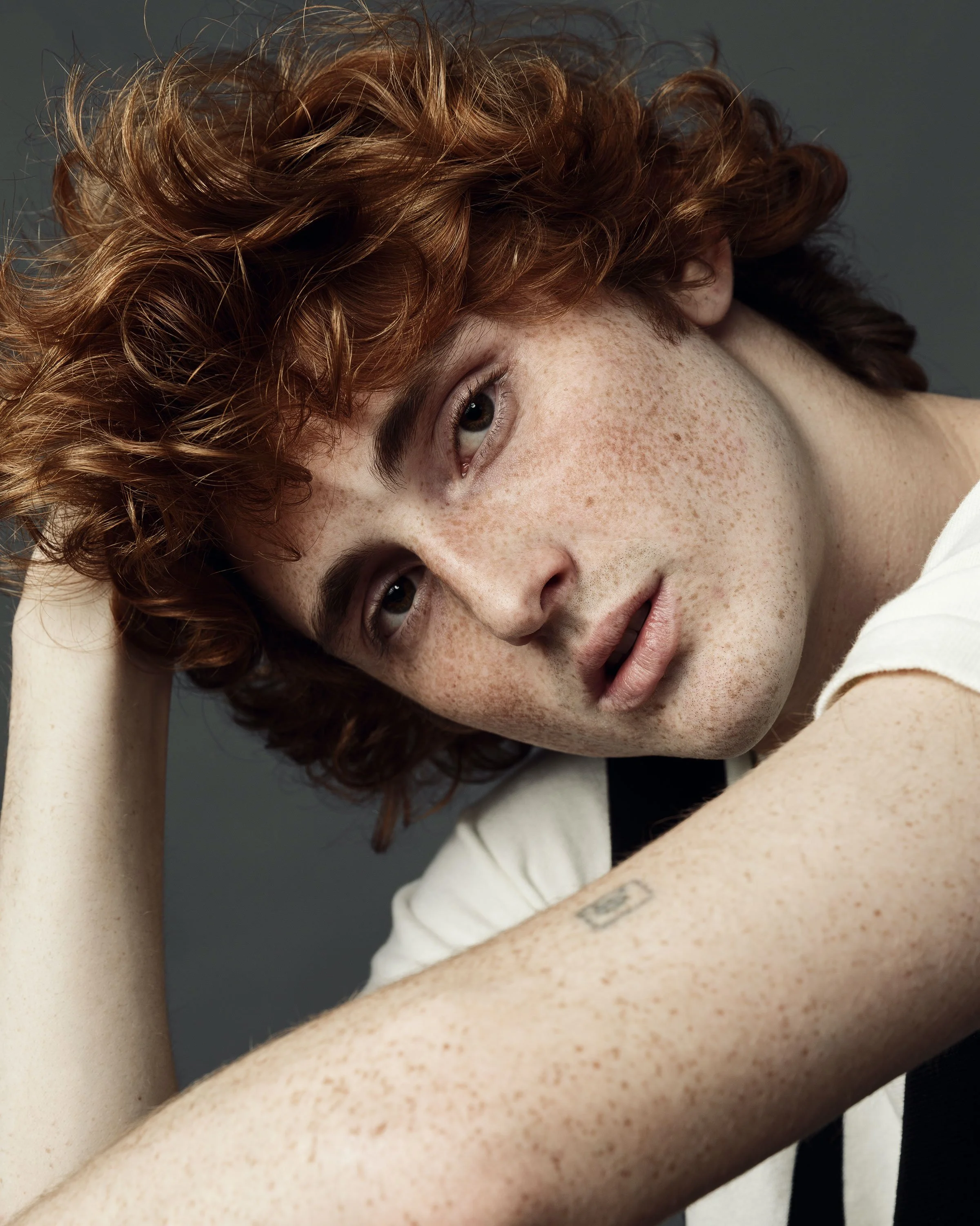 A close-up of a young person with red, curly hair lying down, showing part of their face with freckles, blue eyes, and a faint smile, resting on their arm.
