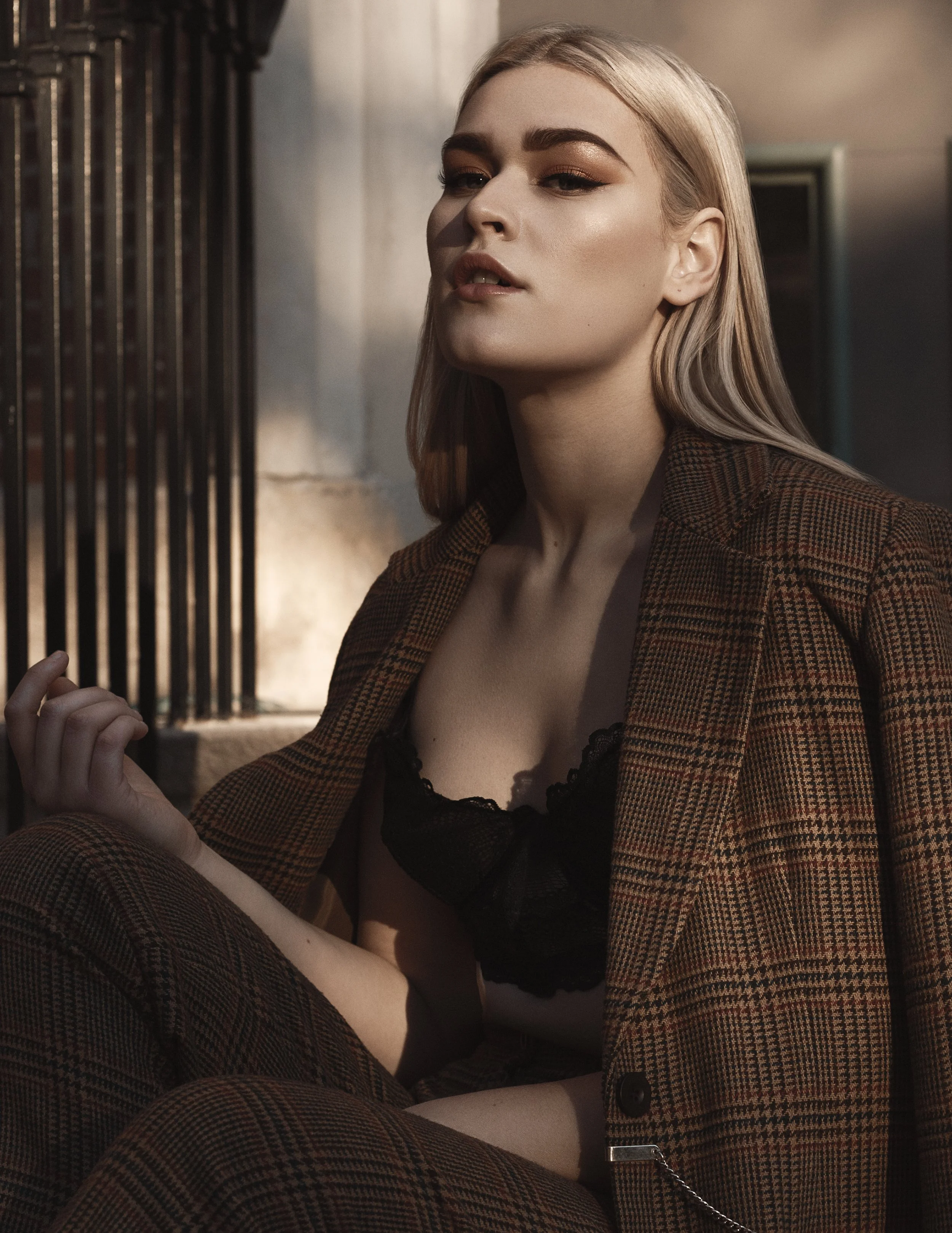 A woman with blonde hair and makeup wearing a plaid blazer and black lace top, sitting against a wall with bars.