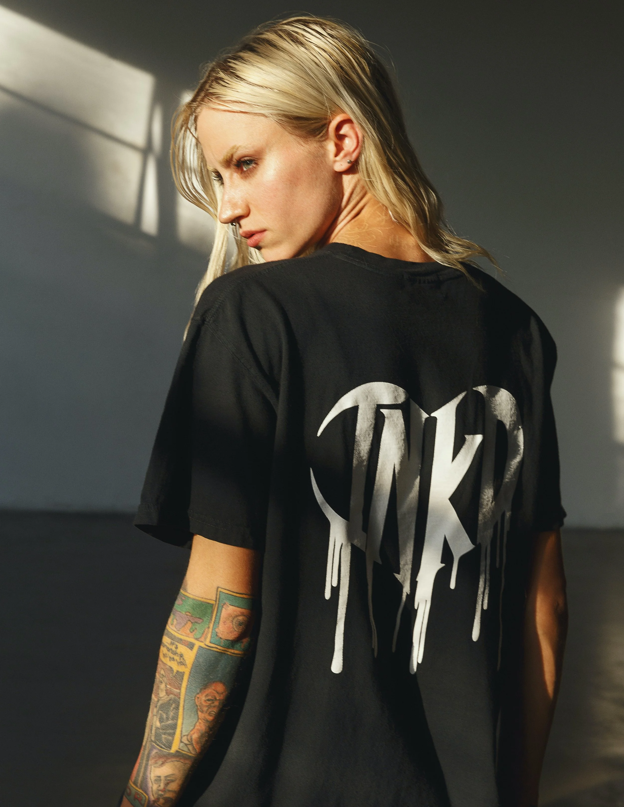 A woman with blonde hair and tattoos on her arm, wearing a black T-shirt, is looking down with sunlight casting shadows on the gray wall behind her.