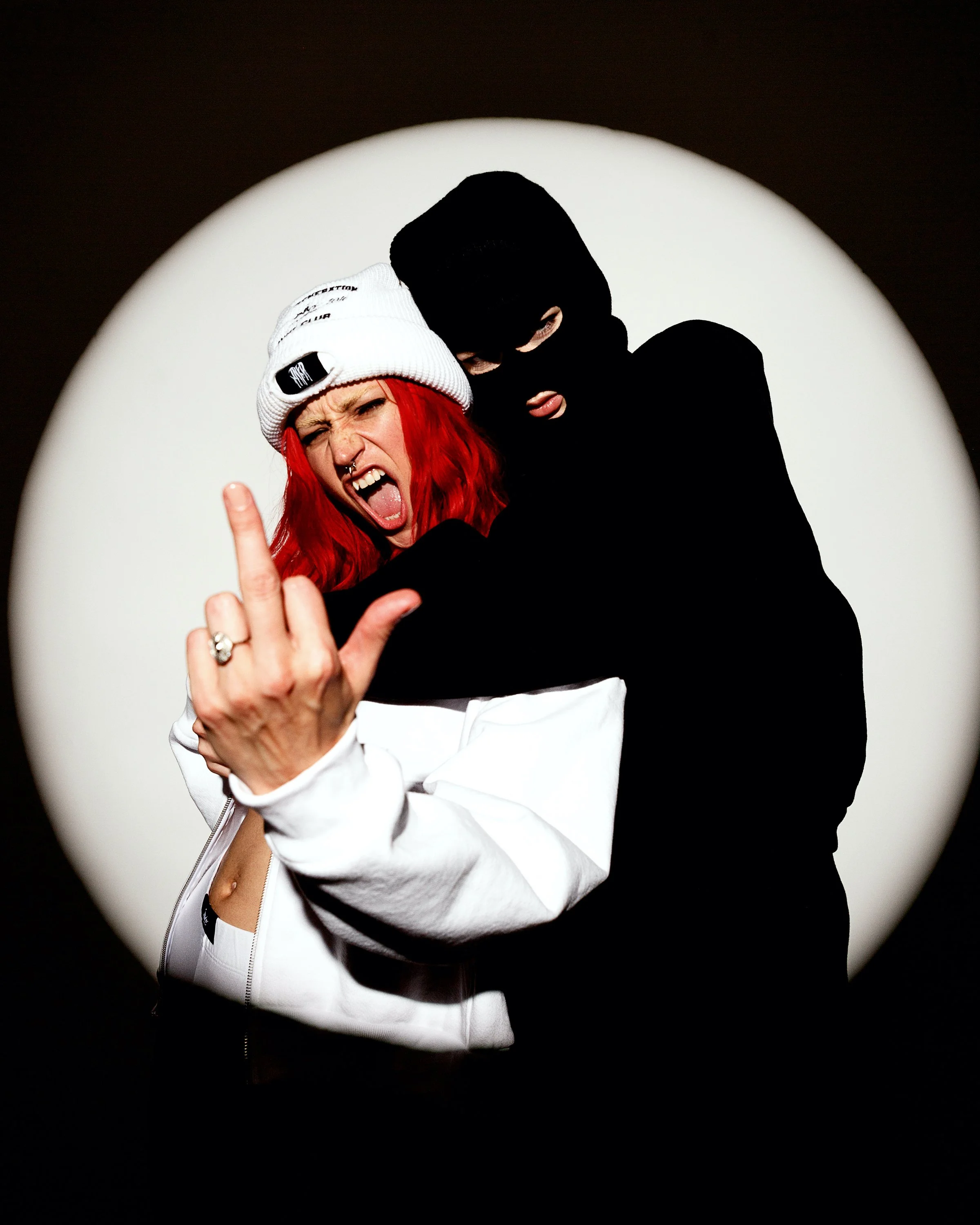 Person with red hair, wearing a white beanie and white jacket, making a rebellious gesture with middle finger raised, alongside a person in a black ski mask and black clothing, posing against a circular spotlight background.