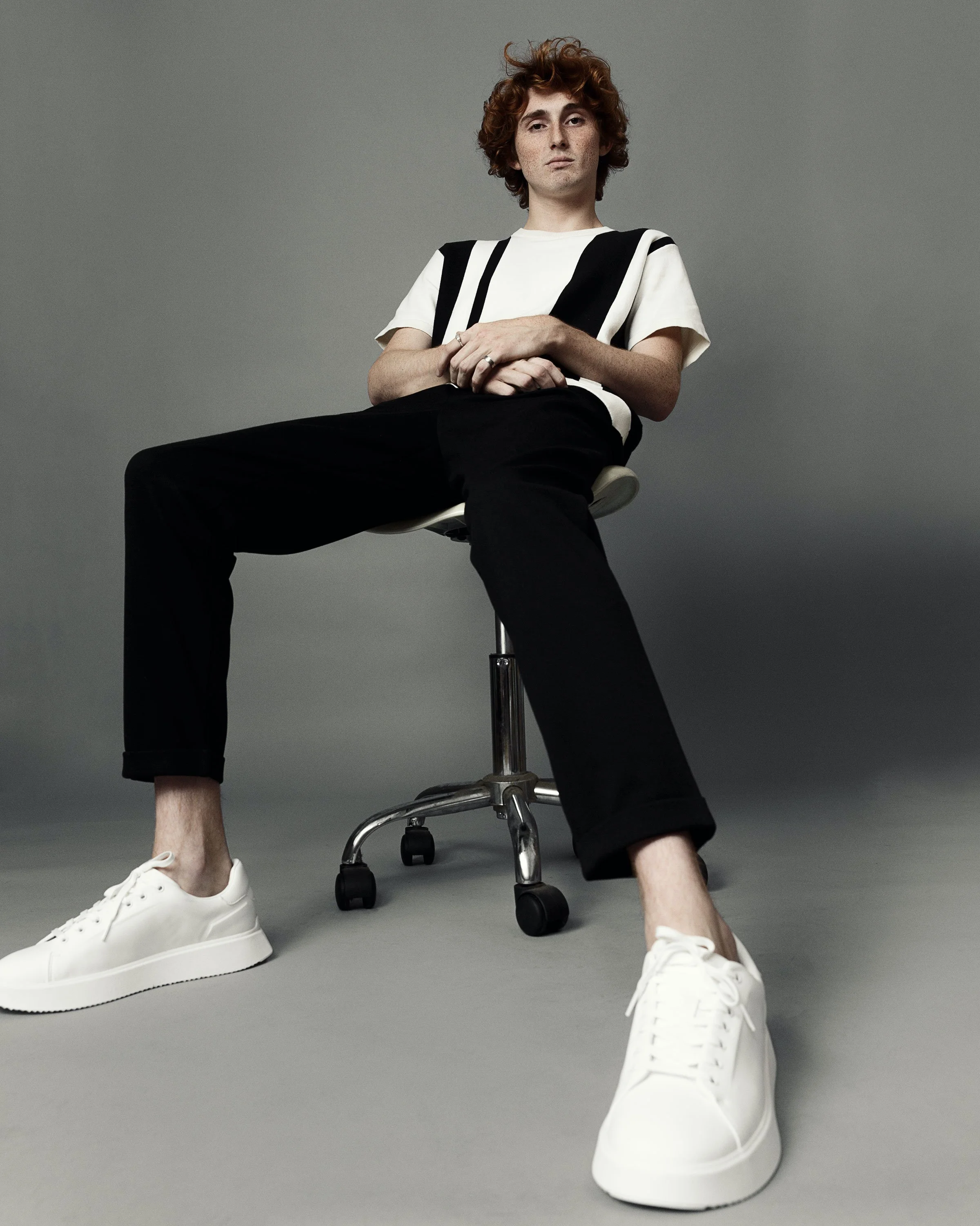 A person with curly brown hair sitting on a black office chair with wheels, wearing a black and white striped shirt, black pants, and white sneakers, against a gray background.