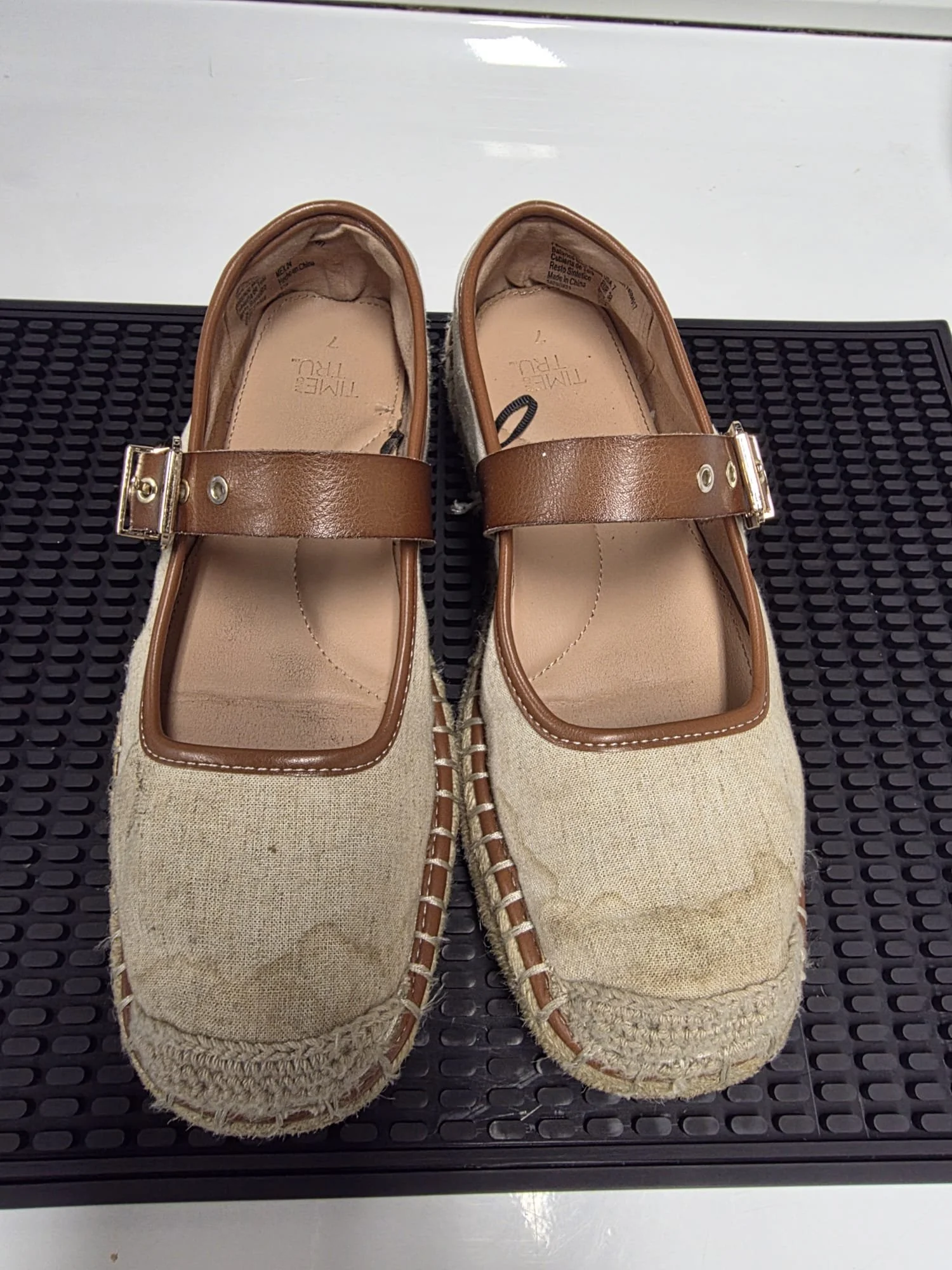 A pair of beige espadrille shoes with brown leather straps and metal buckle, placed on a black rubber mat.