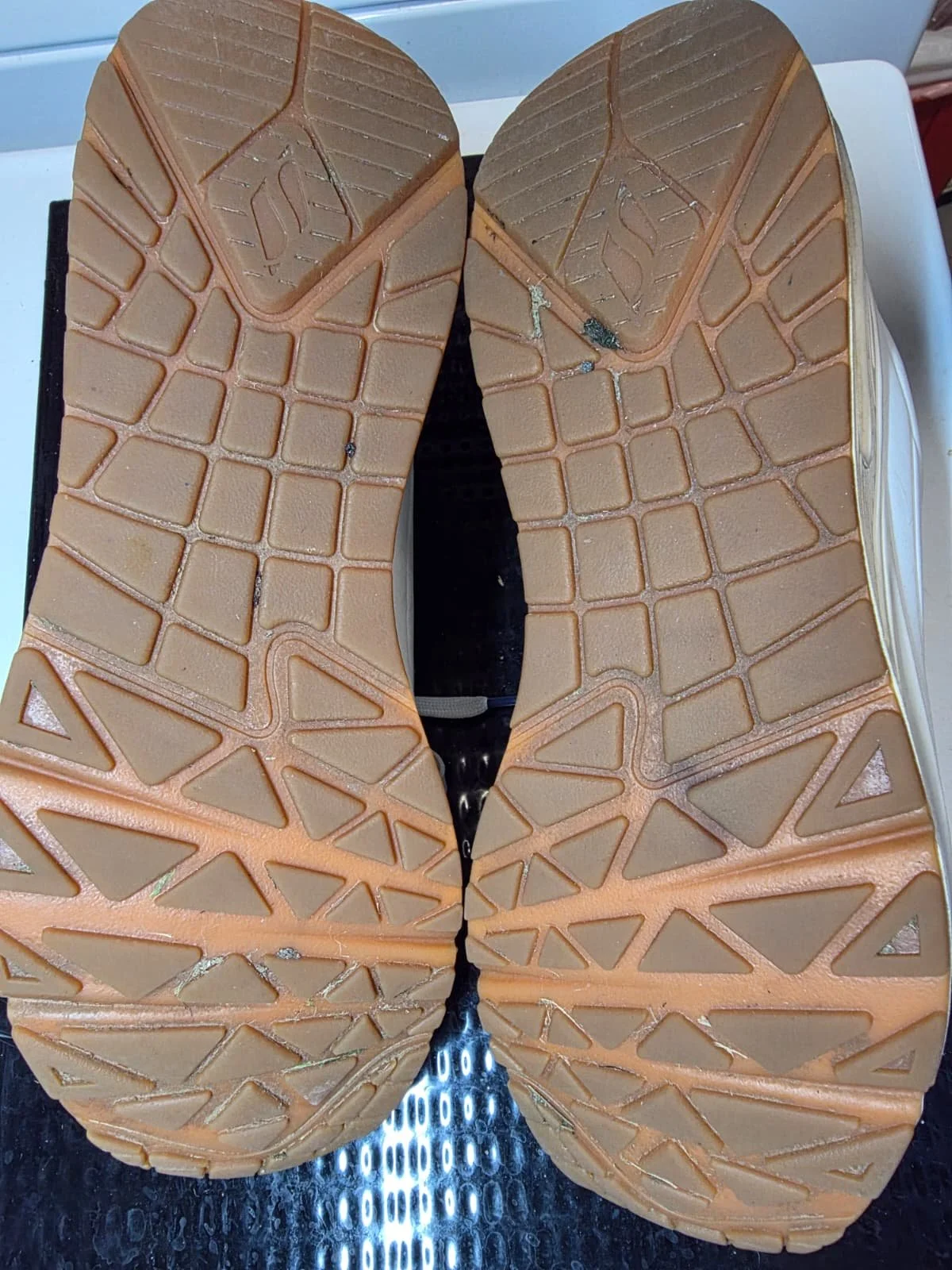 The image shows the soles of a pair of beige sports shoes with a geometric pattern on the bottom, placed on a metal surface.
